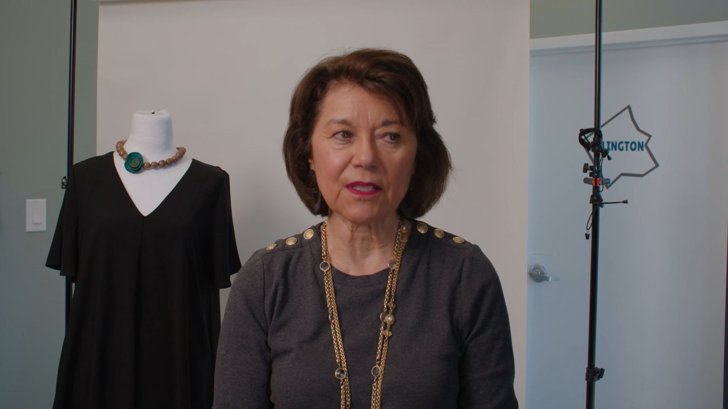 A professional woman wearing a black blazer and necklace stands against a white wall in an office setting.