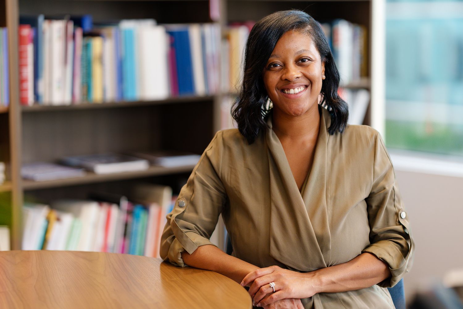 Professional in a beige blazer sits at desk with bookshelf background in bright office setting.