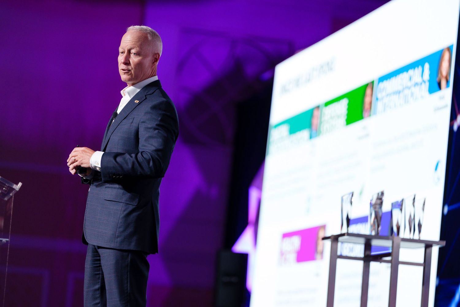 Business speaker in dark suit presents colorful digital slides at corporate conference event on stage.