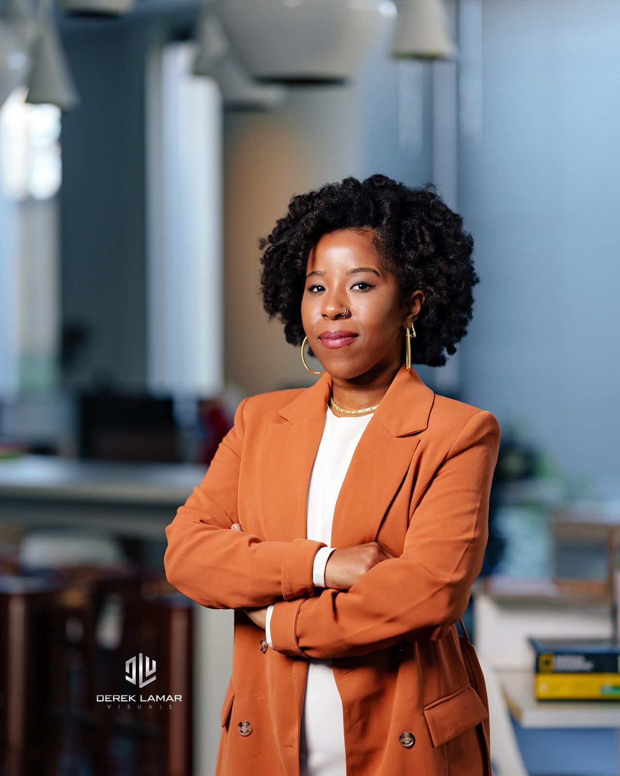 Professional wearing an orange blazer and white top stands confidently with arms crossed in modern office setting.