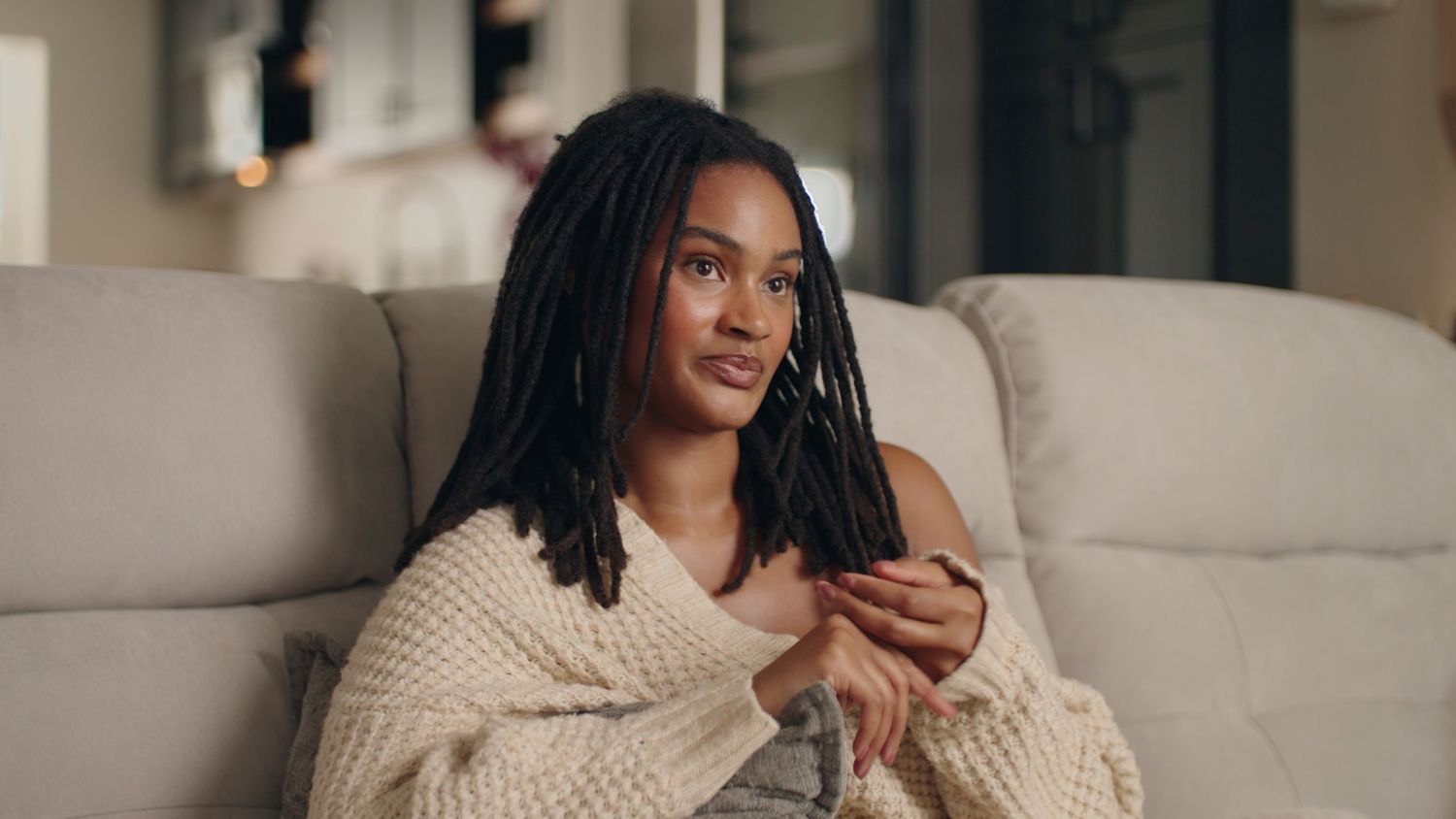 Young woman with braided hair relaxing on couch wrapped in cozy knit sweater in modern living room.