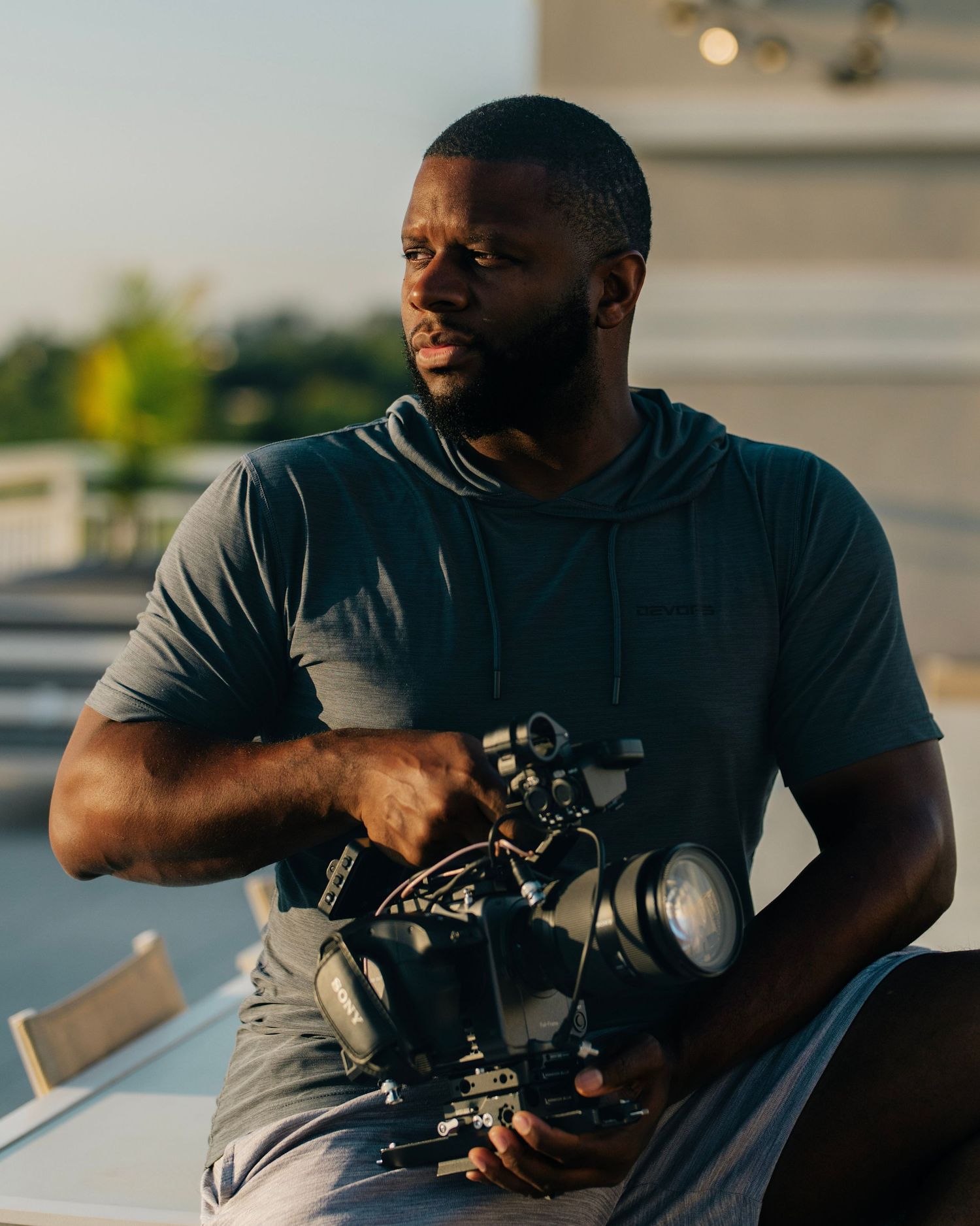 A man in a gray hoodie holds a professional camera on a rooftop during golden hour.