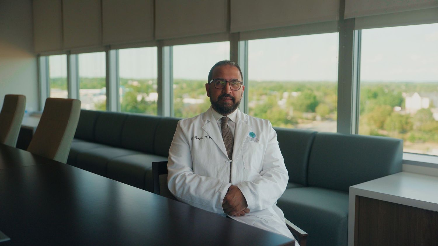 Medical professional in white coat stands confidently in modern hospital corridor with panoramic windows.
