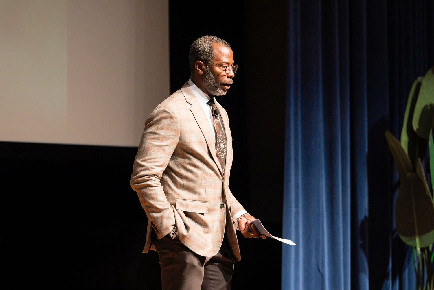 Speaker wearing a light beige suit gestures energetically while presenting on stage with blue curtain backdrop.