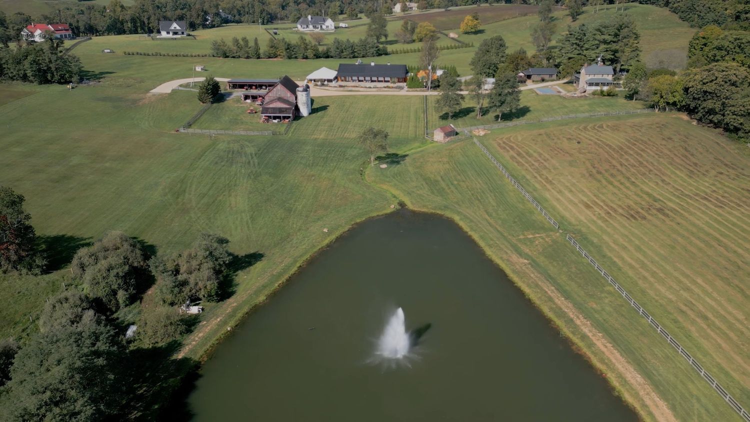 Aerial view of a rural property with a fountain spraying in a pond surrounded by farmland and buildings.