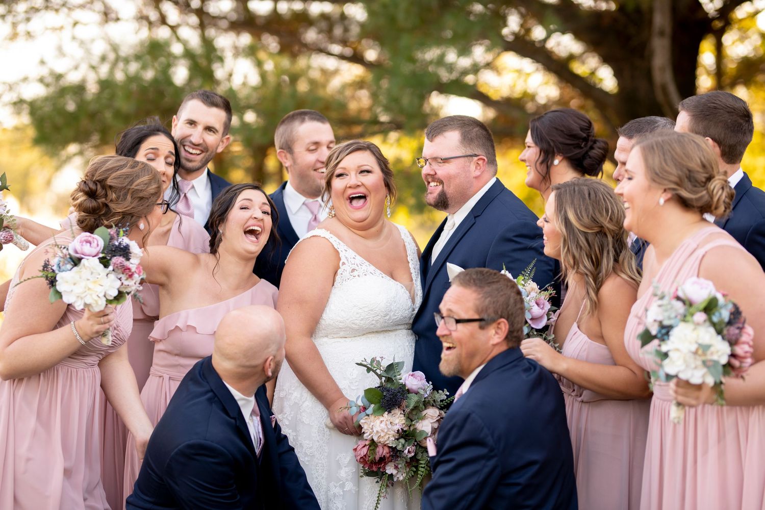 Wedding party laughs together outdoors in pink and navy attire while holding white floral bouquets at sunset.