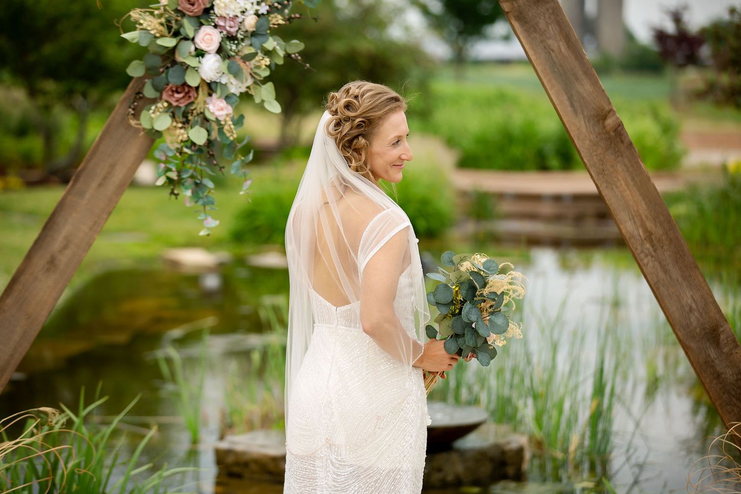 A bride in a white lace dress holds a bouquet while standing by a wooden arch with floral decorations near a pond.