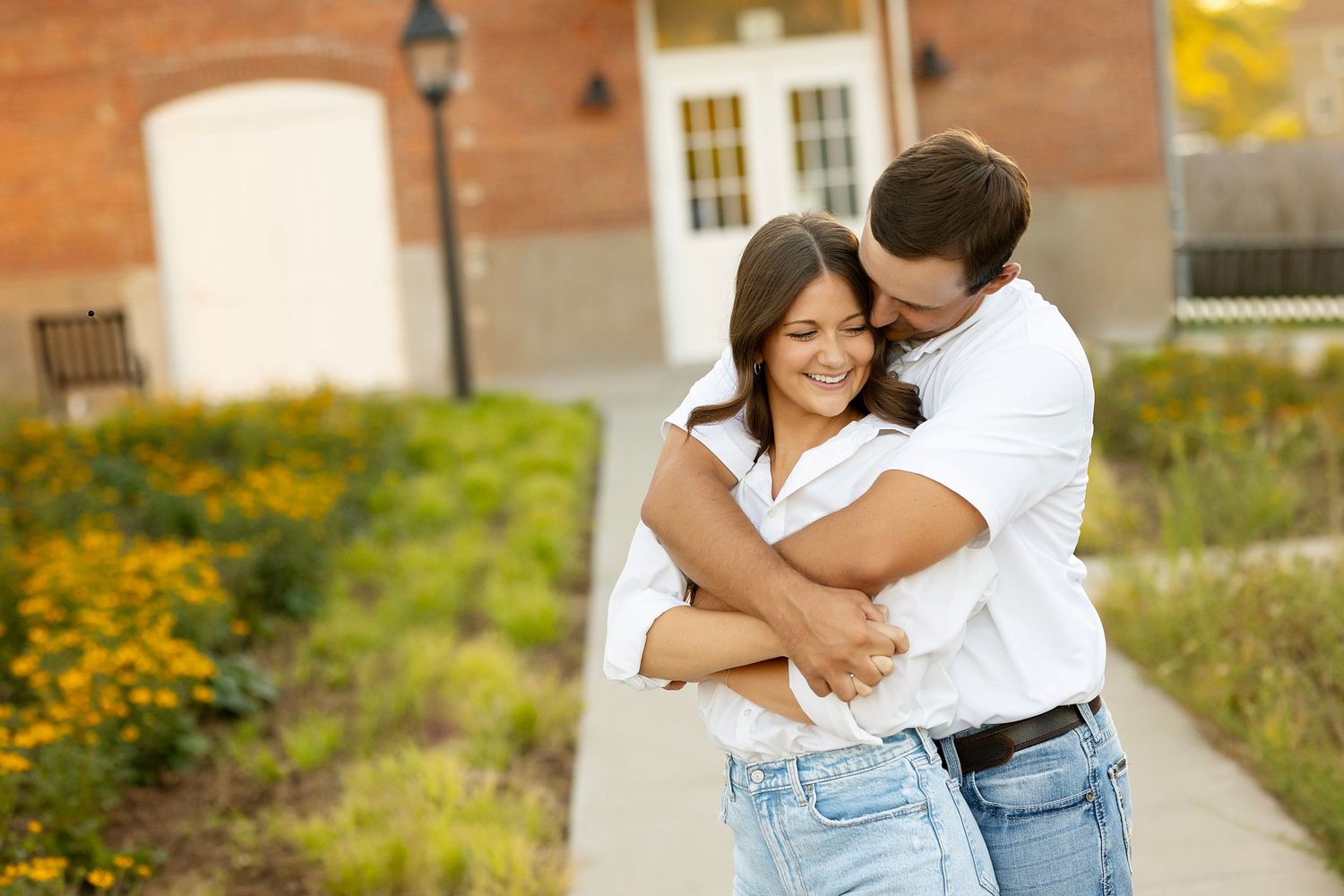 A couple embraces lovingly on a walkway with yellow wildflowers and rustic buildings in the background.