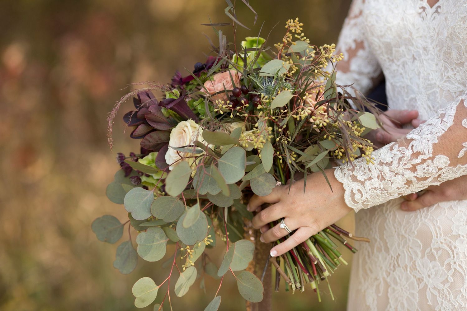 Beautiful bridal bouquet with eucalyptus leaves, purple flowers and blush roses held against lace wedding dress.