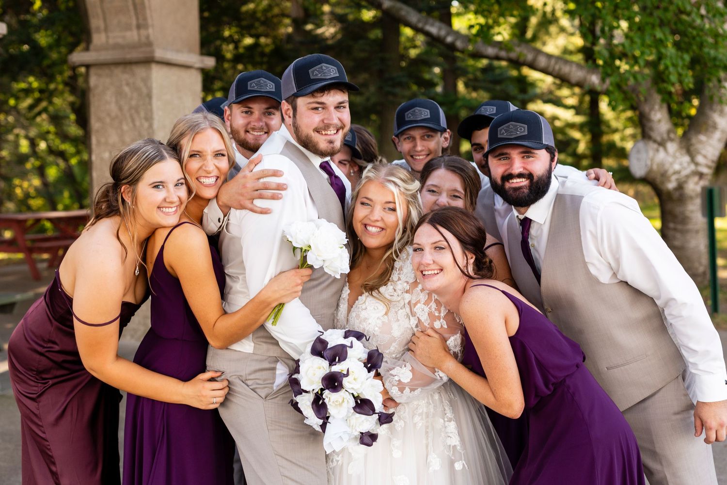 Wedding party poses together outdoors in burgundy and grey attire with bouquets for a playful group photo.