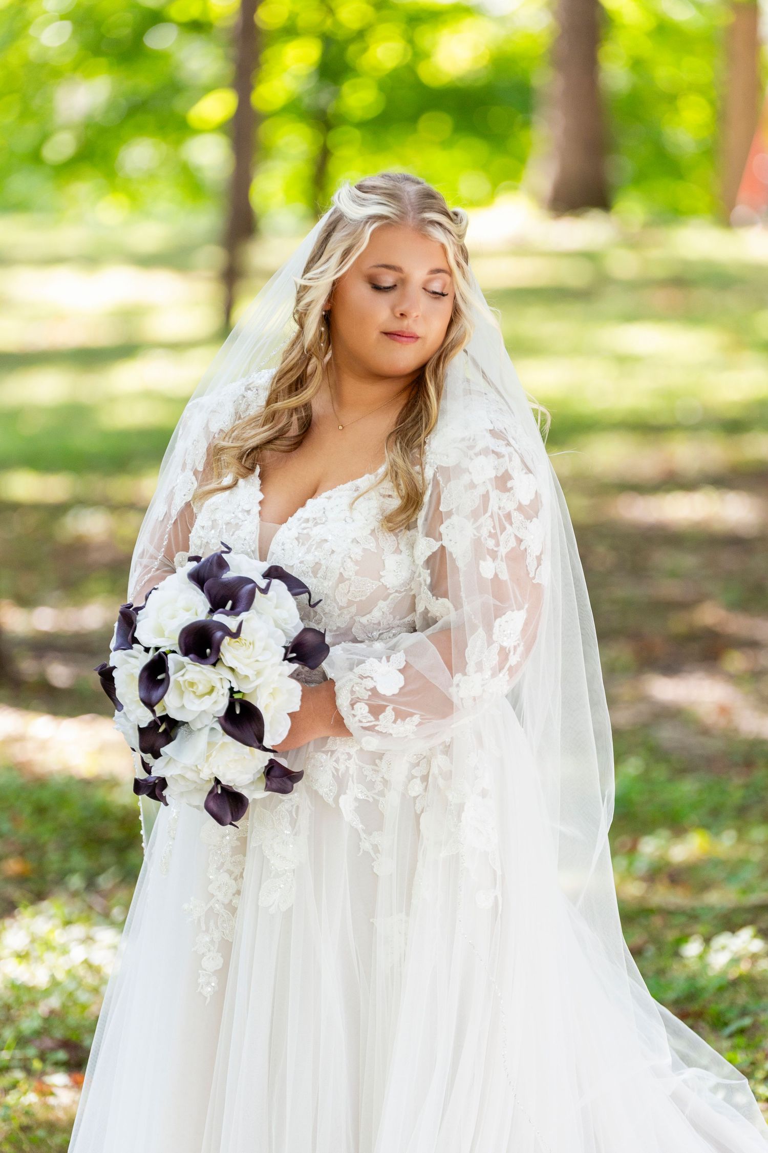 A bride in a white lace wedding dress with long sleeves and veil holds a stunning purple and white bouquet in a sunlit garden.