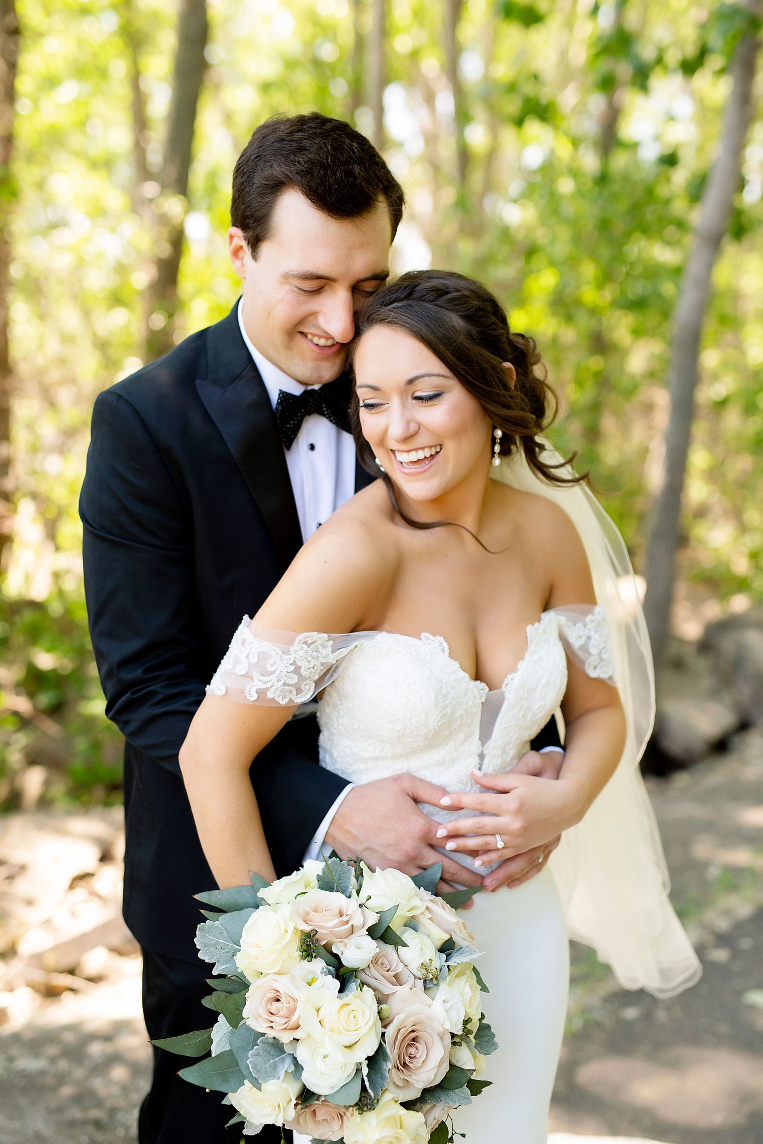 Newlyweds share an intimate moment outdoors while holding a bouquet of blush roses and white flowers with dusty miller.
