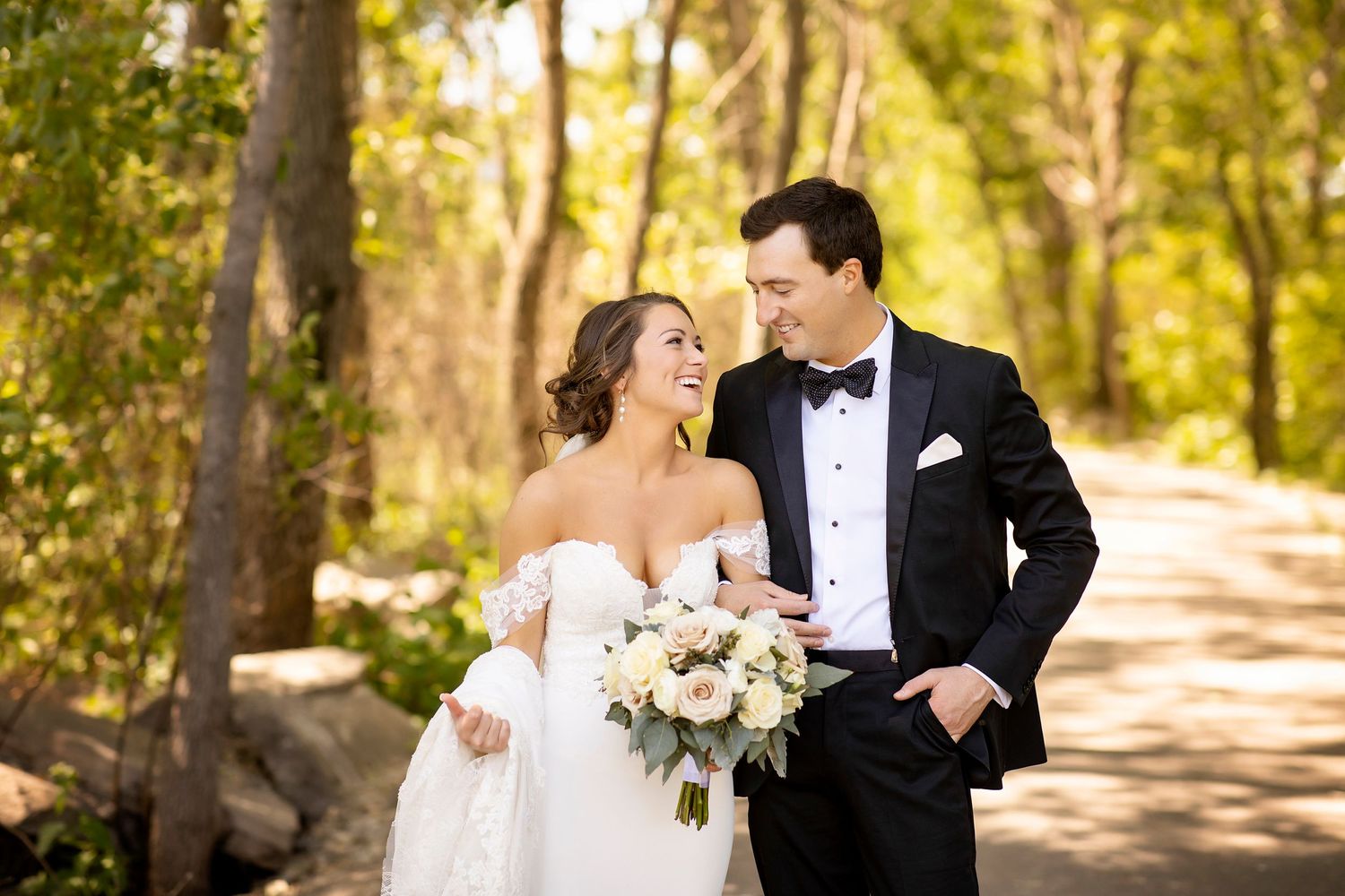 A couple shares a joyful moment on a sunlit forest path, with the bride holding a blush and white bouquet.