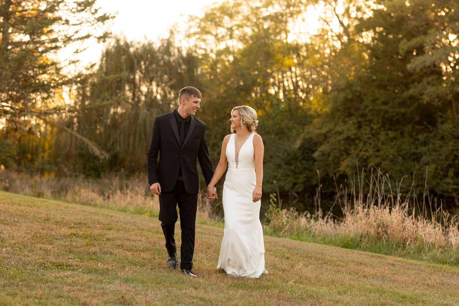 A couple walks hand in hand through a golden sunlit field at sunset wearing formal wedding attire.