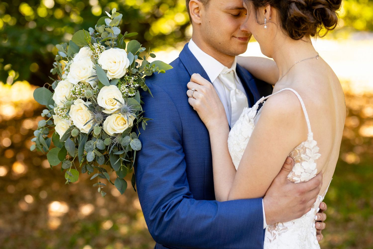 A couple embraces at their outdoor wedding, with a beautiful white rose and eucalyptus bouquet in view.