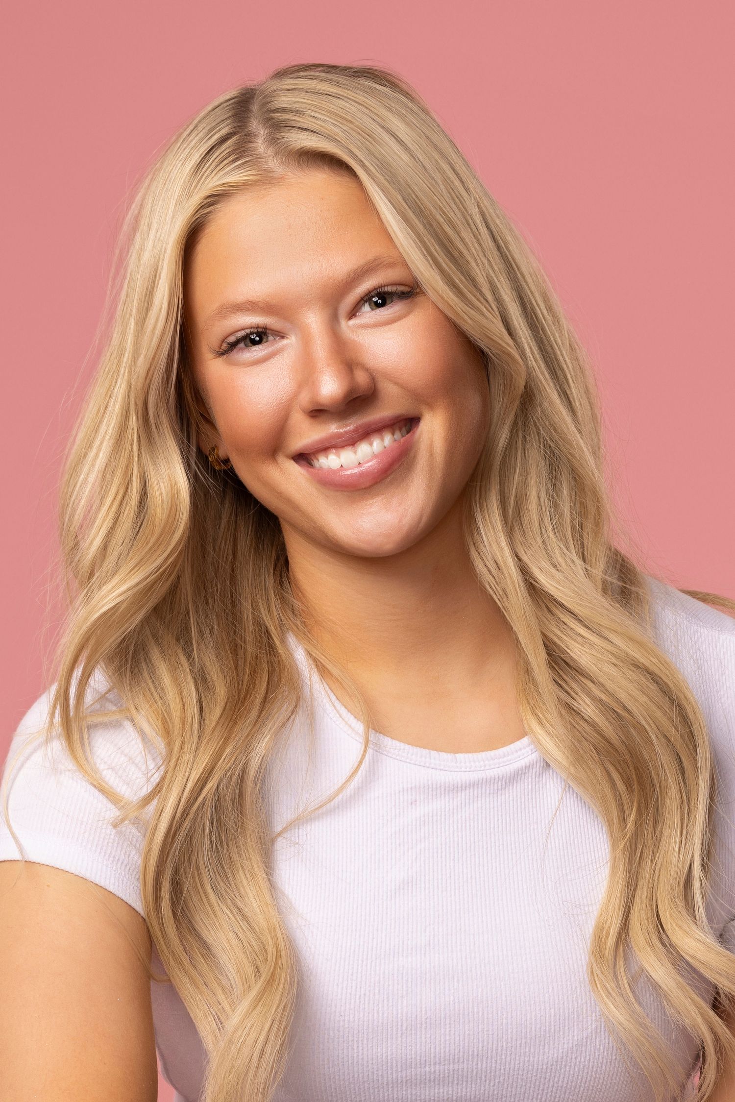 Portrait of a person with wavy blonde hair wearing a white t-shirt against a pink background, showing a bright smile.