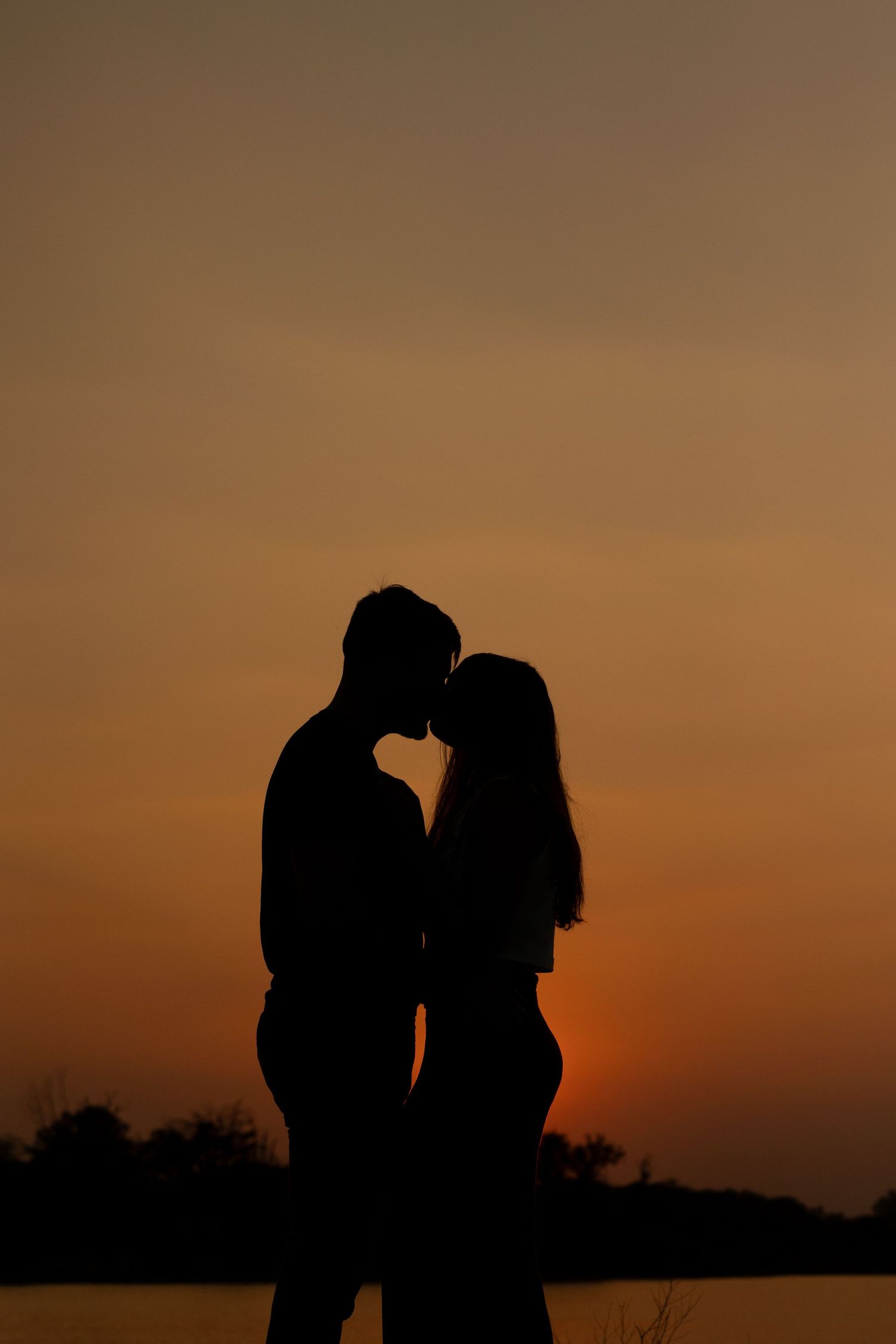 A romantic silhouette of a couple sharing an intimate moment against a warm orange sunset sky.