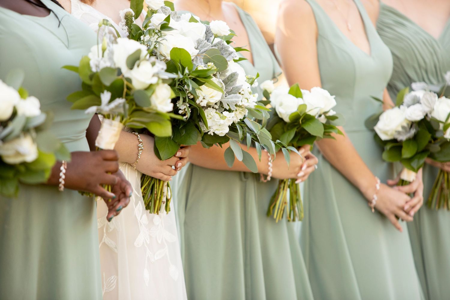 Bridesmaids in sage green dresses hold elegant white and green floral bouquets at wedding ceremony.