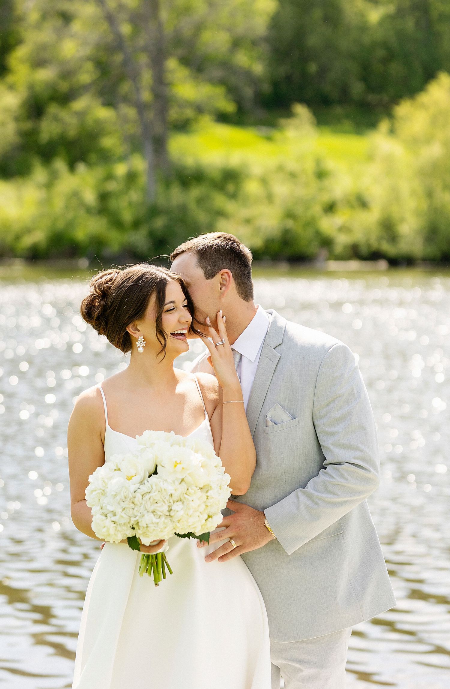 Romantic wedding couple sharing an intimate moment by a sparkling lake while holding a bouquet of white flowers.