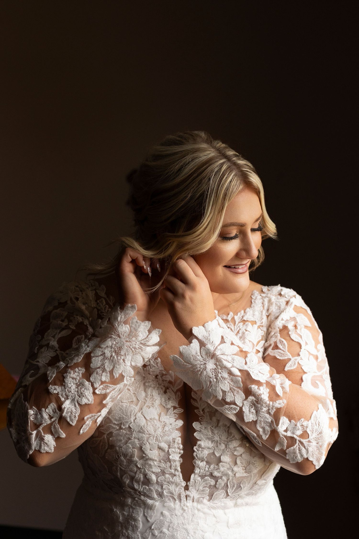 A person wearing an elegant white lace wedding gown with long sleeves puts on jewelry in a dimly lit room.