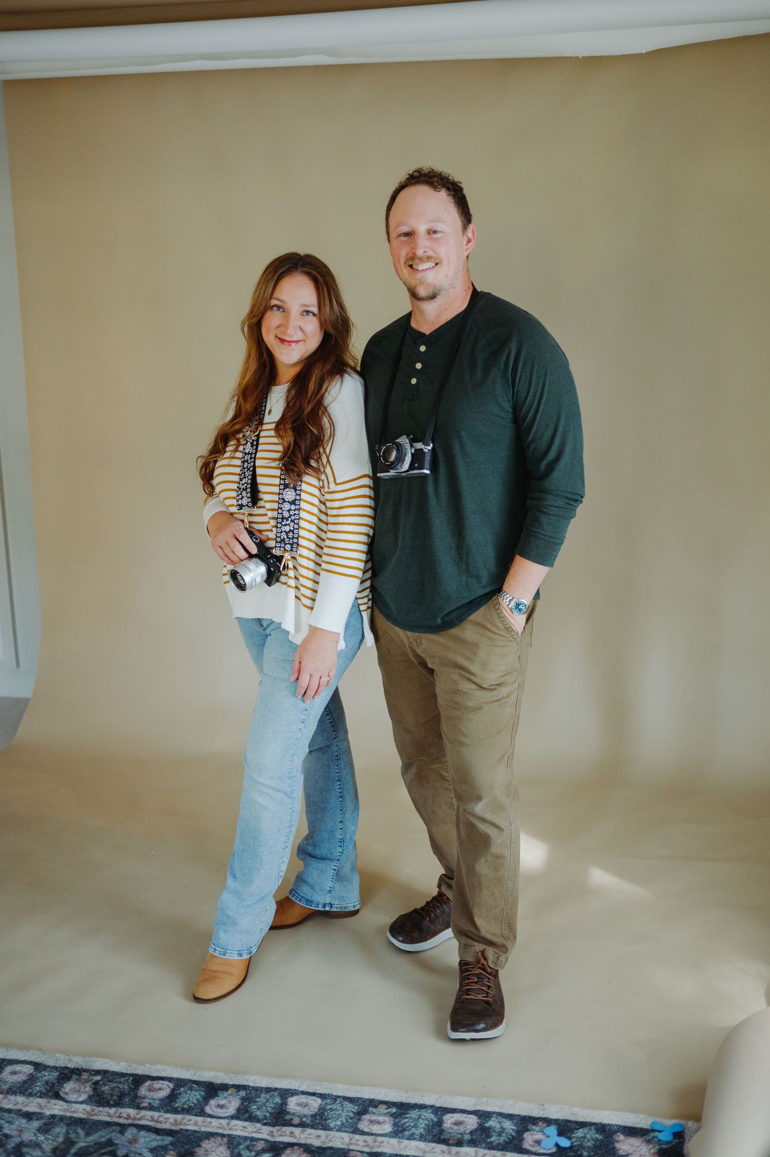 A photographer couple poses playfully together in a casual indoor setting with light beige walls and decorative border trim.