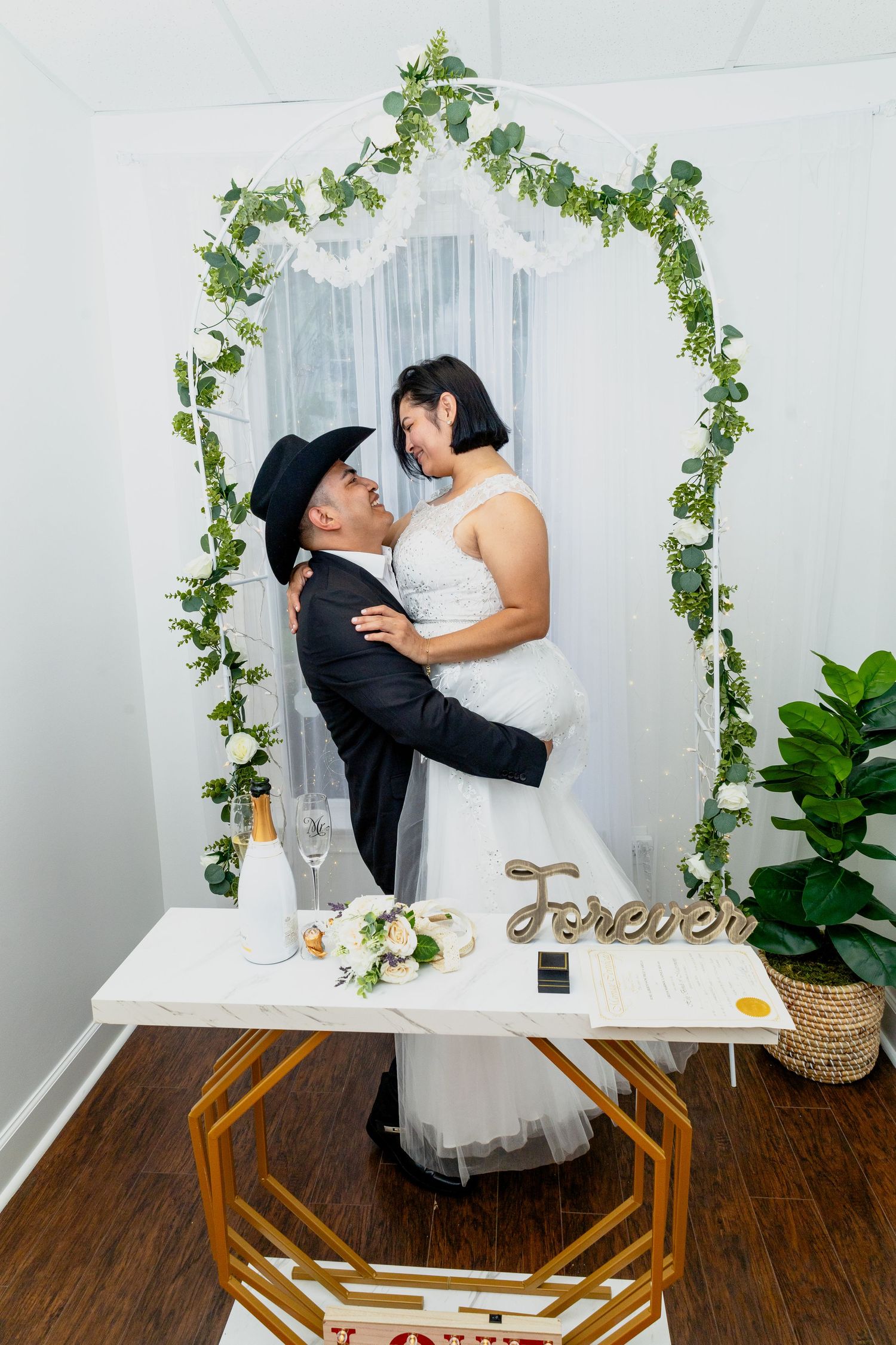 Wedding couple posing under a floral arch with greenery and white curtain backdrop.