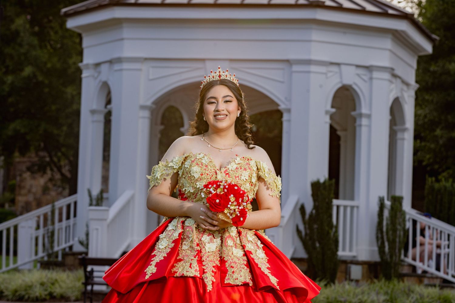 An elegant red and gold quinceañera dress with intricate lace details photographed by a white gazebo.