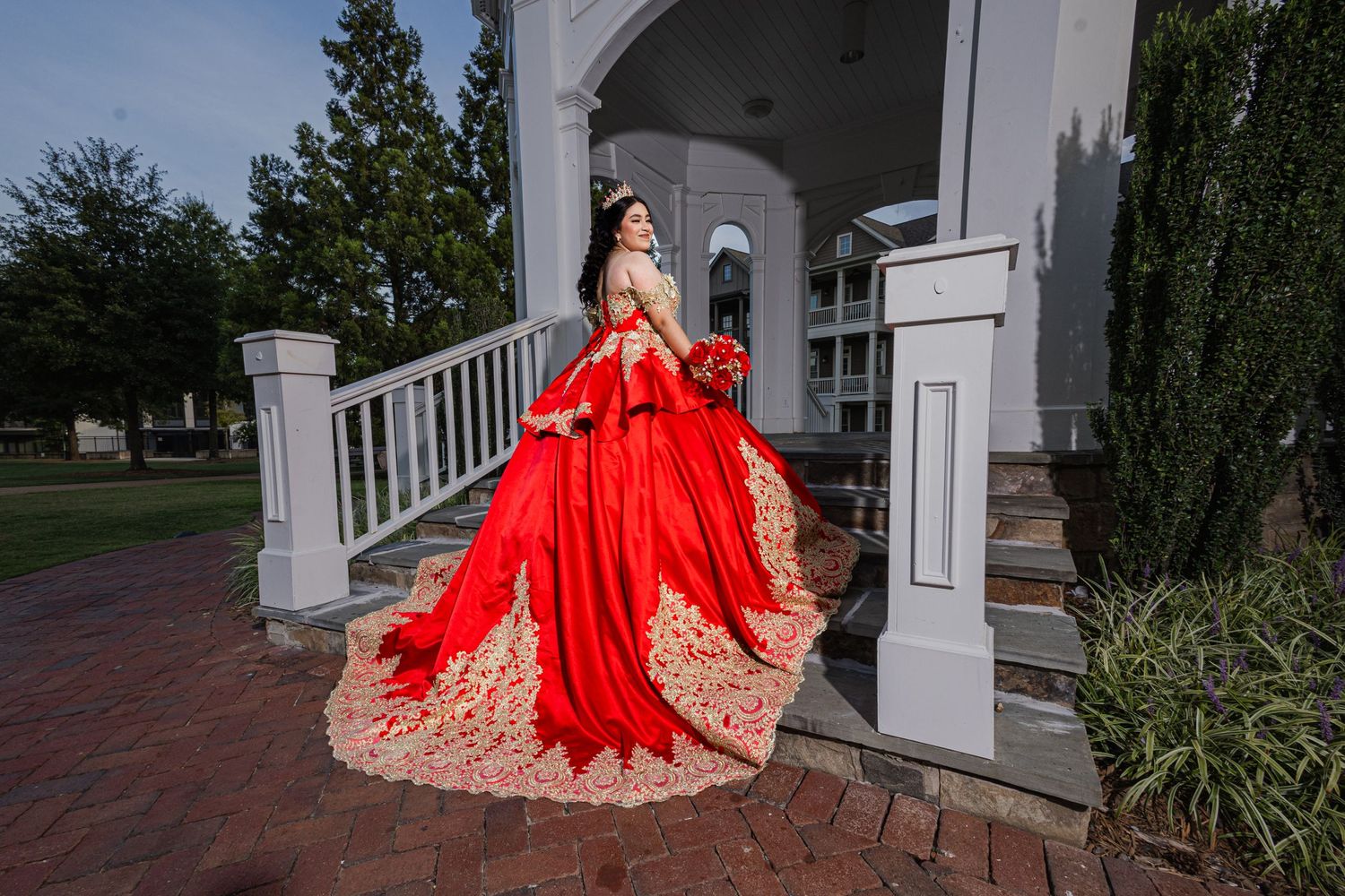 Elegant red and gold formal dress displayed against white colonial-style architecture.