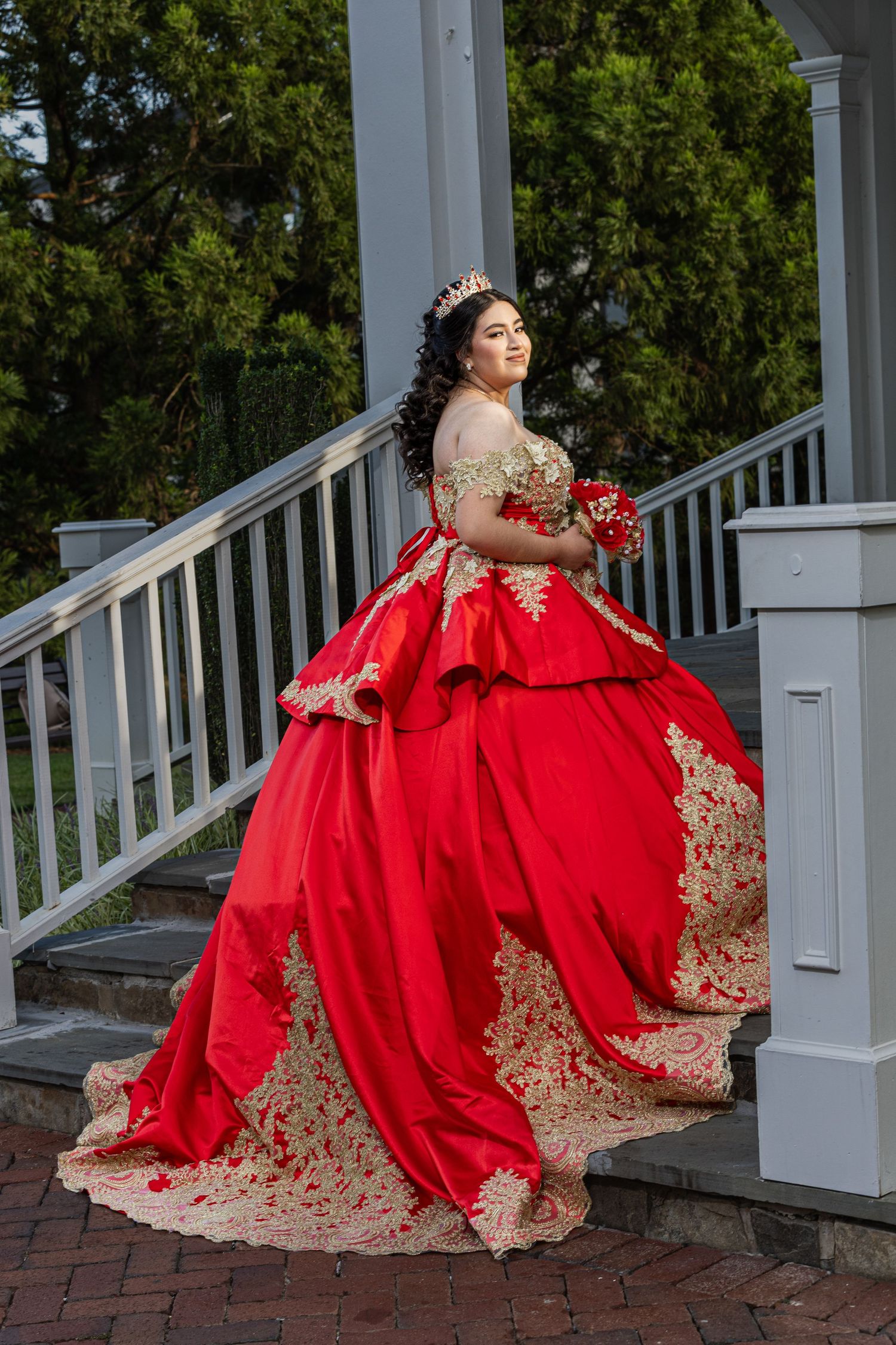 Stunning red ball gown with gold embellishments photographed on brick patio near white columns.
