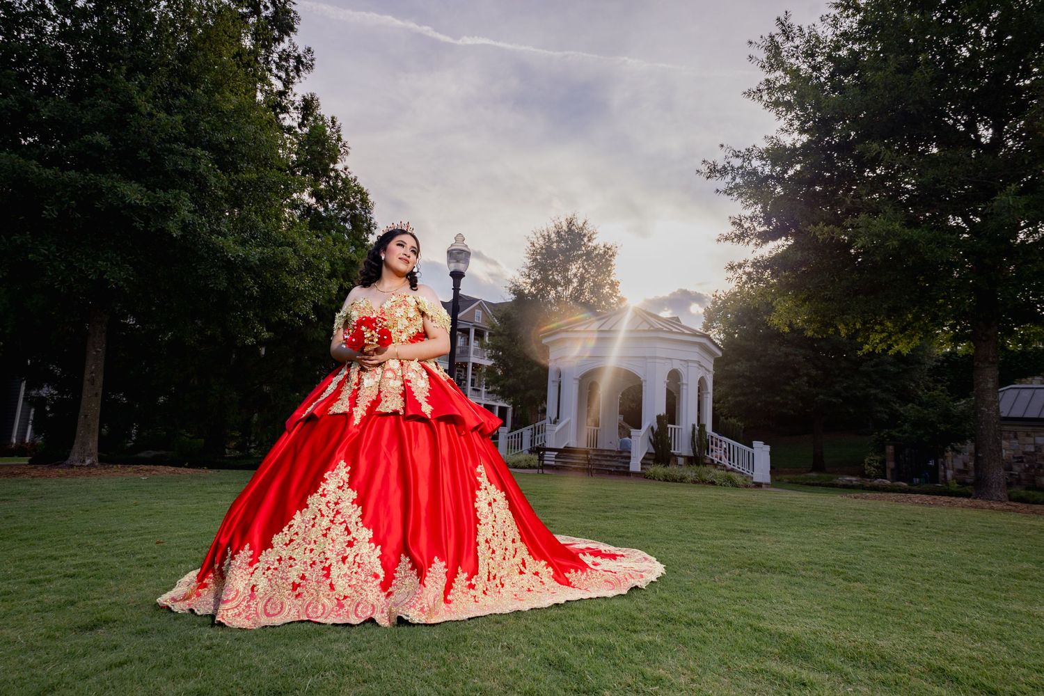 A stunning red and gold ball gown photographed in front of a white gazebo during golden hour.