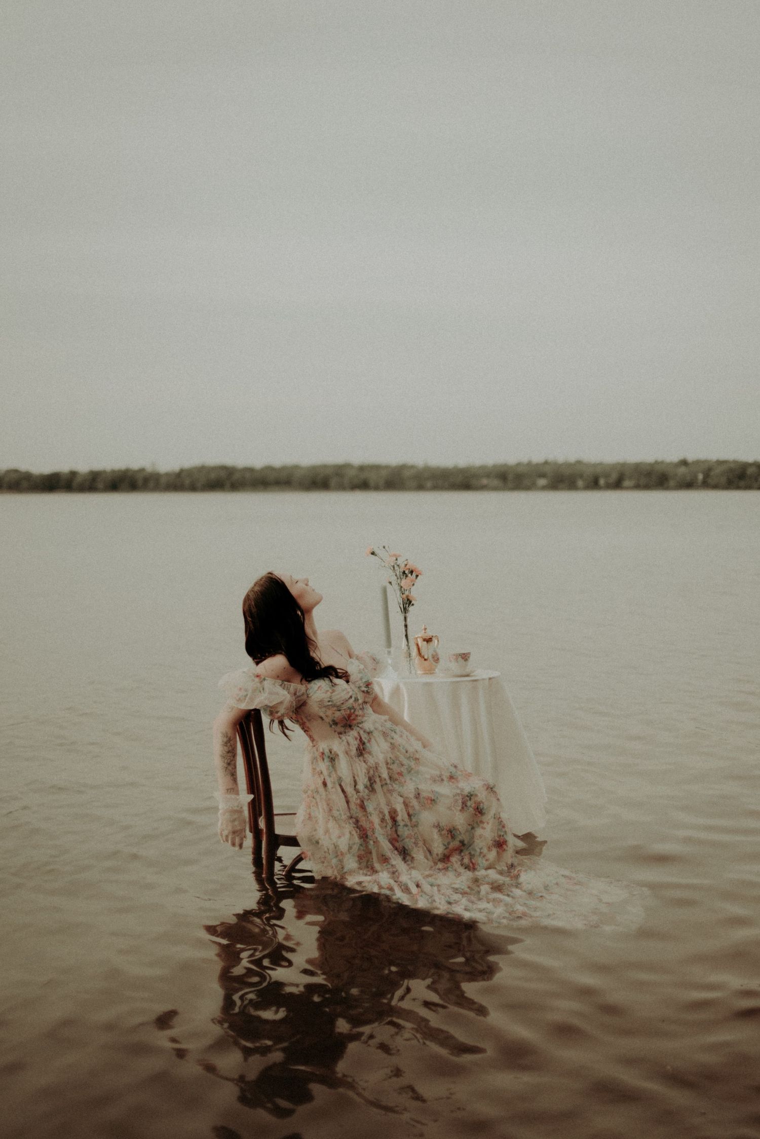 Artistic portrait of someone in a flowing dress seated in water with a small table and floral centerpiece.