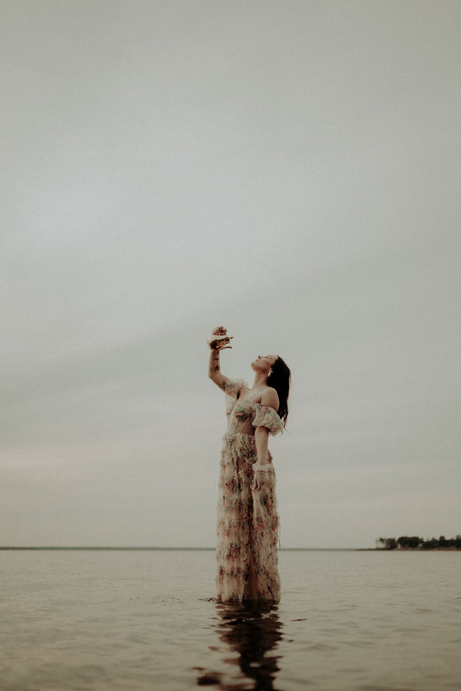 Artistic portrait of someone in a flowing dress standing in water with arms raised.