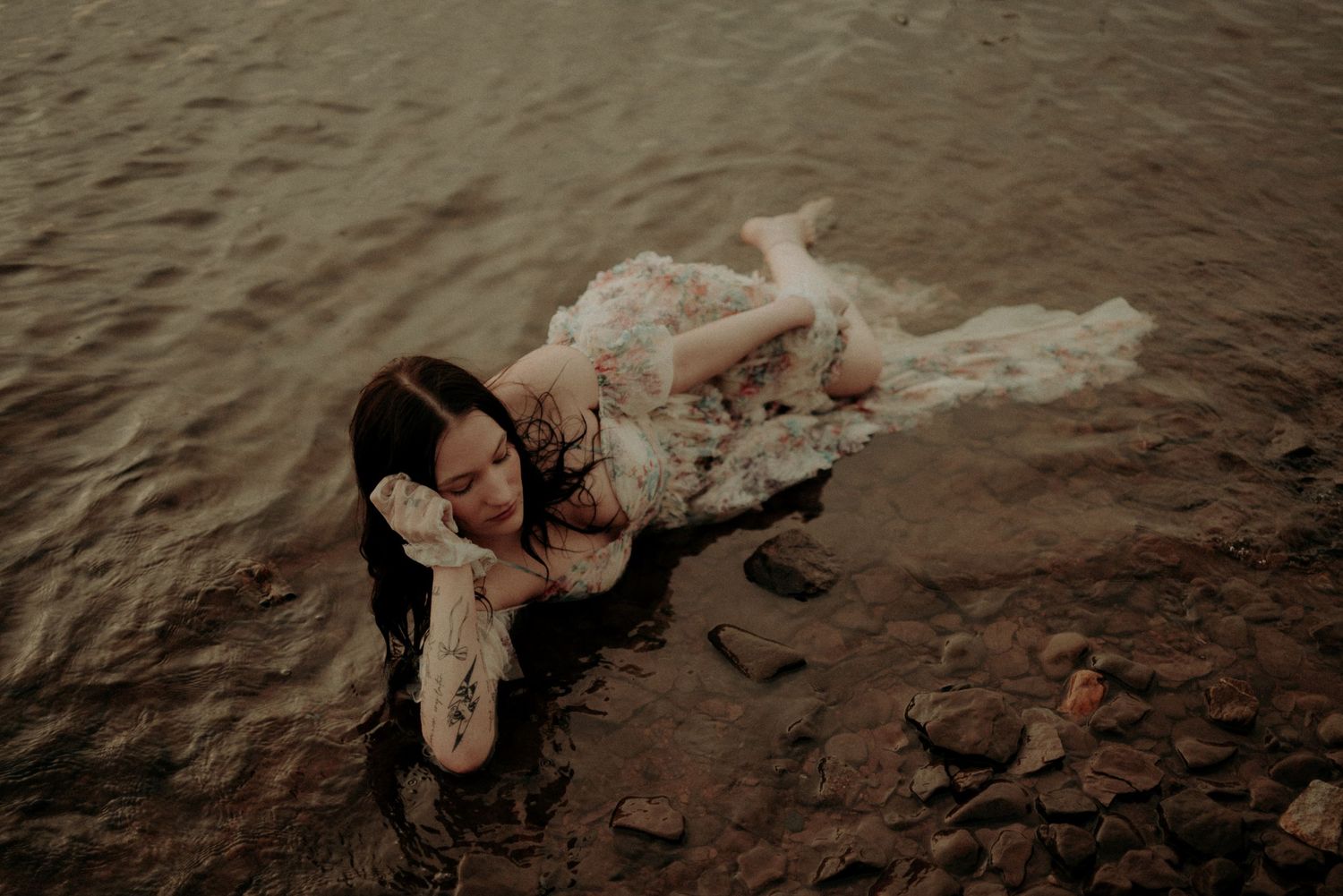 Atmospheric photo of a person in lace dress lying in shallow rocky waters at twilight.