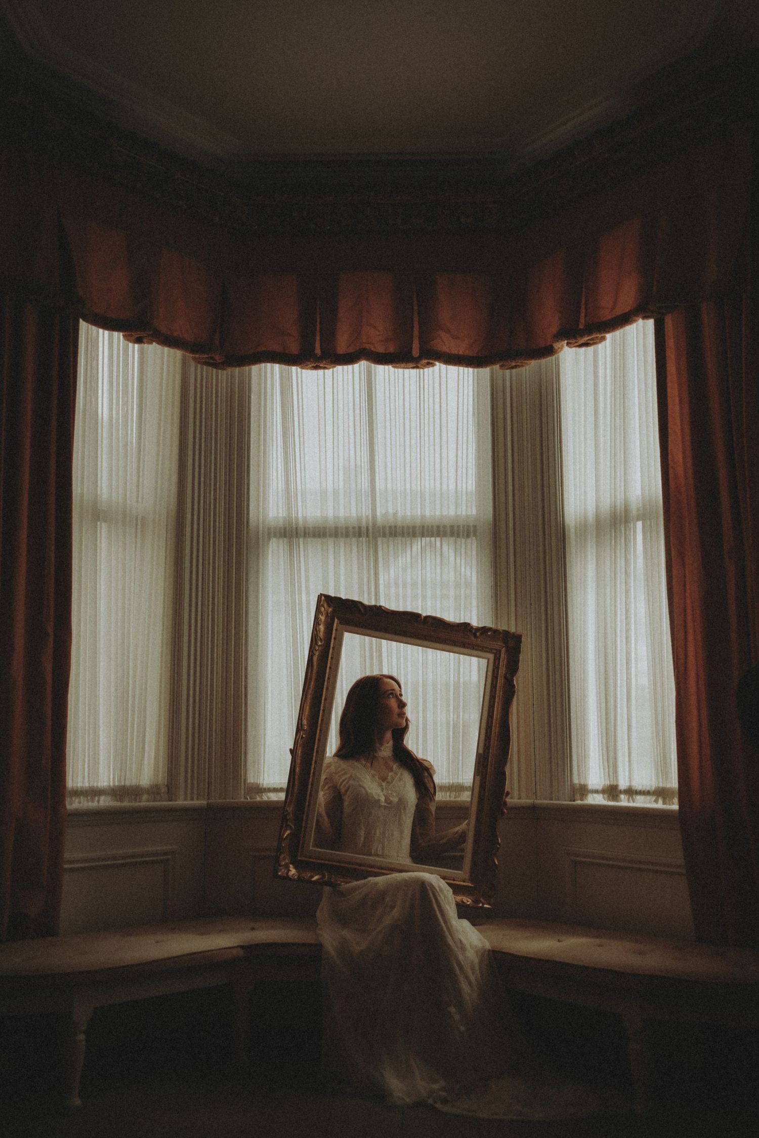 Atmospheric bridal portrait through antique frame in window alcove with burgundy valance.