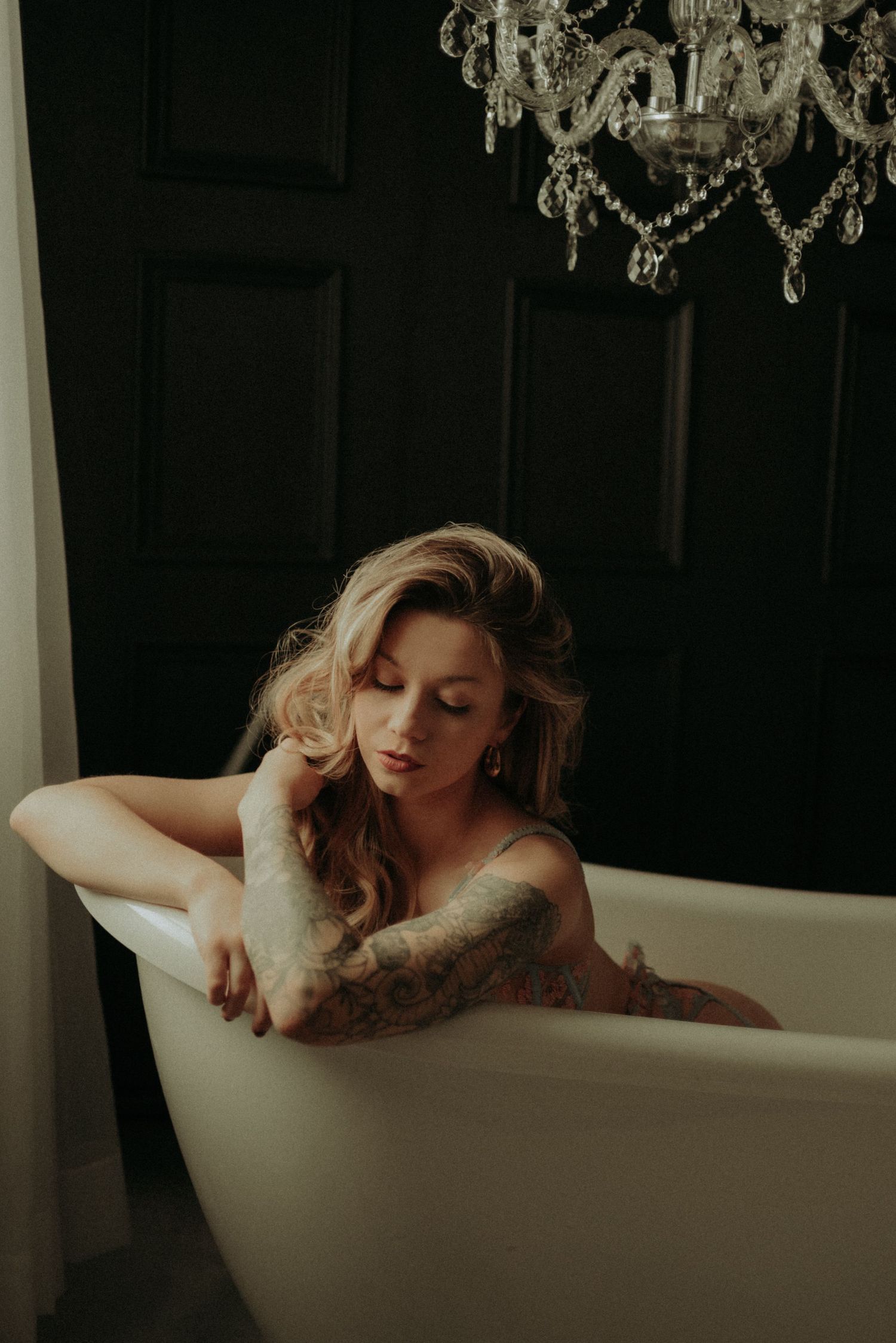 Tasteful boudoir photo features a crystal chandelier above white clawfoot bathtub in moody dark setting.