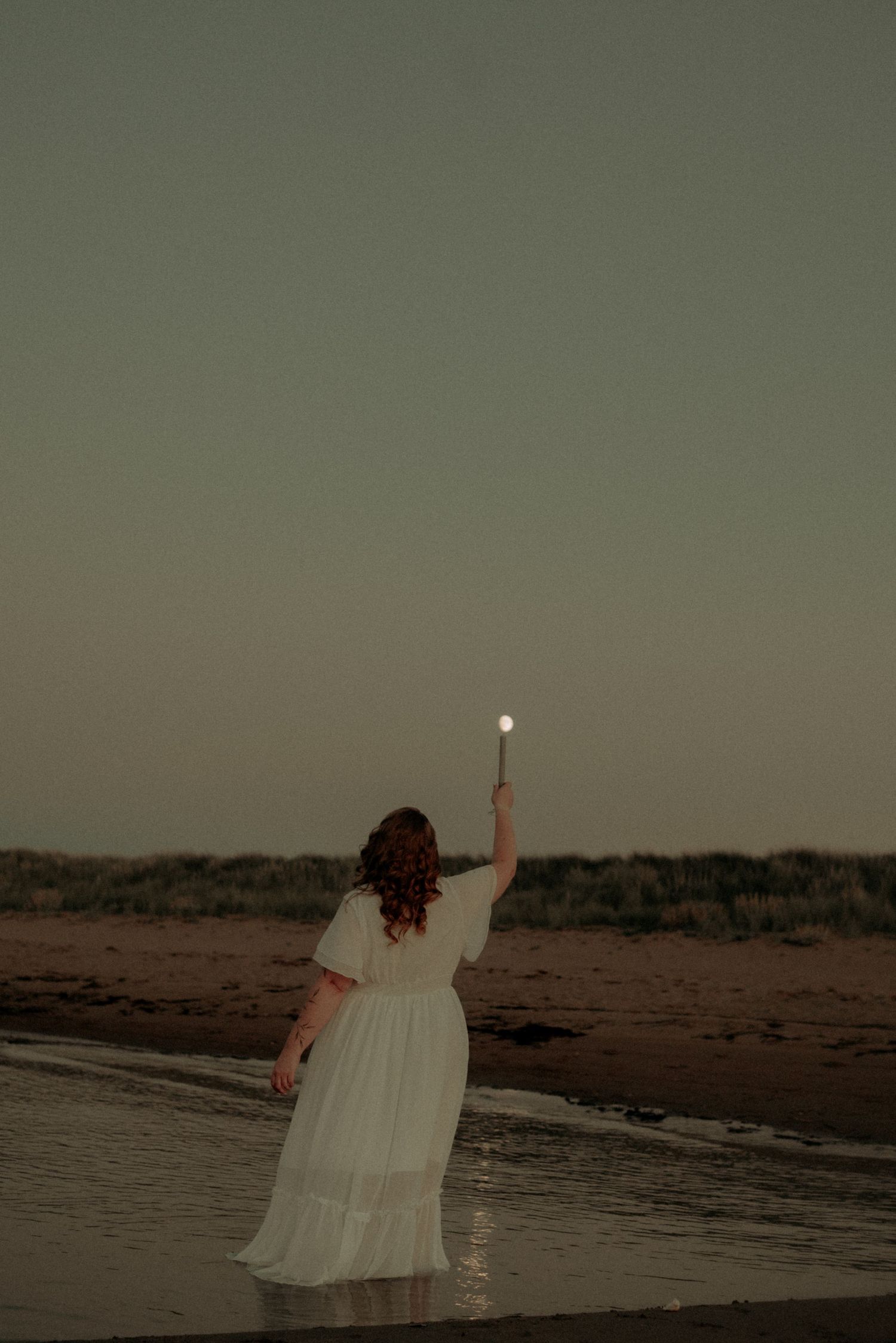 A silhouette of a person in a white dress stands on a beach at dusk against a moody sky.