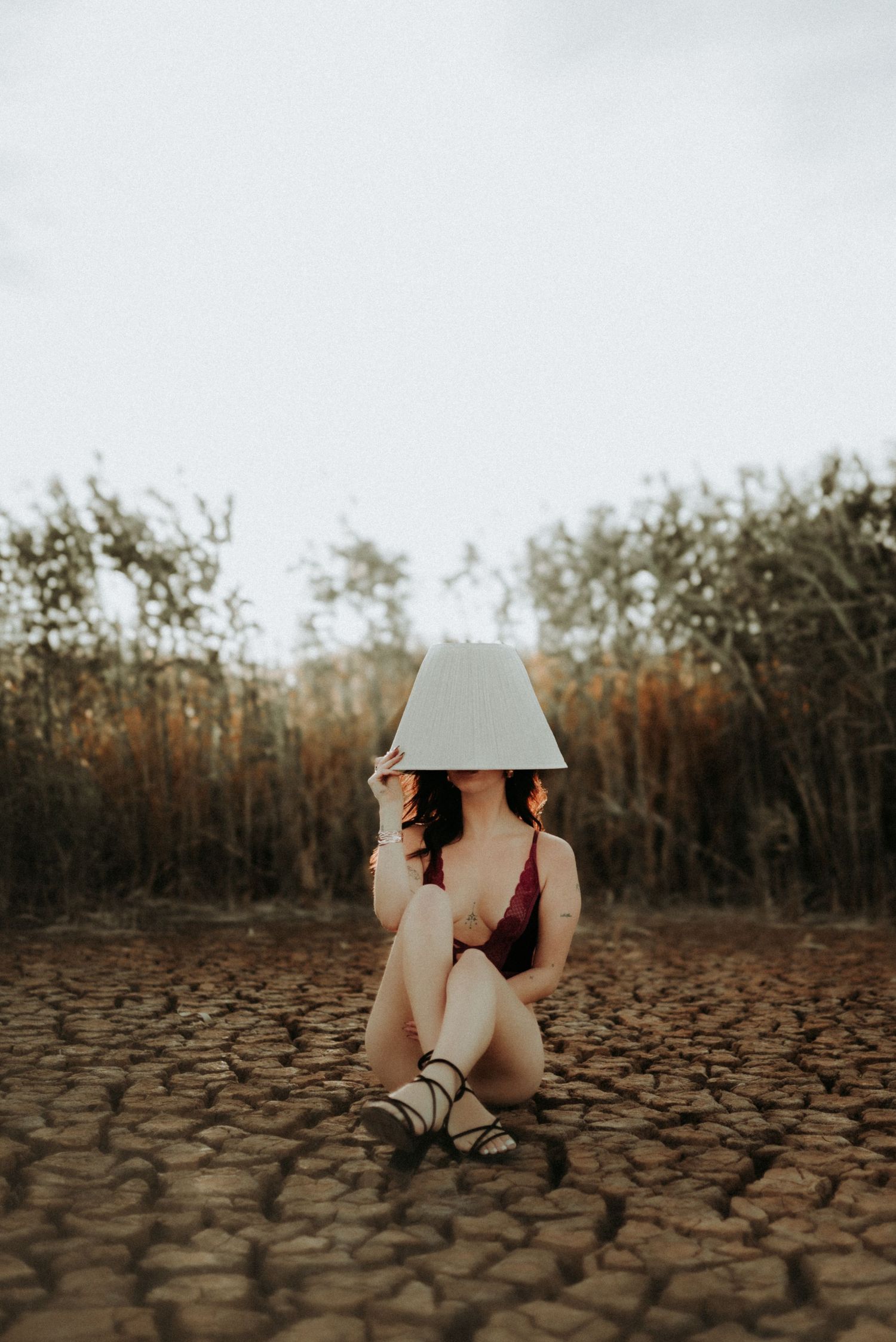 Figure in a hat poses on cracked dry earth surrounded by tall grass in an artistic nature photo.