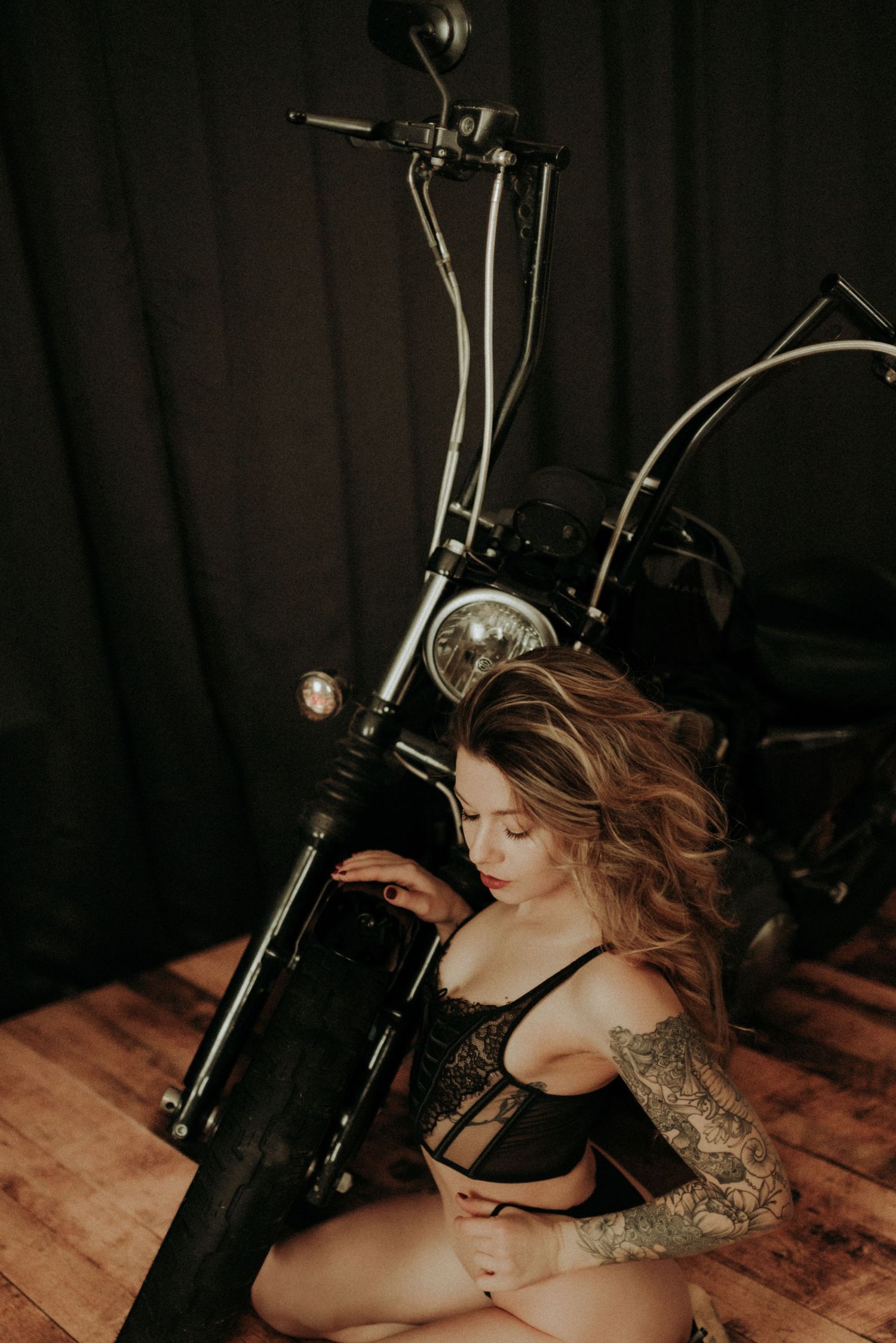 Dark boudoir photography featuring a vintage motorcycle on wooden floor against black backdrop.