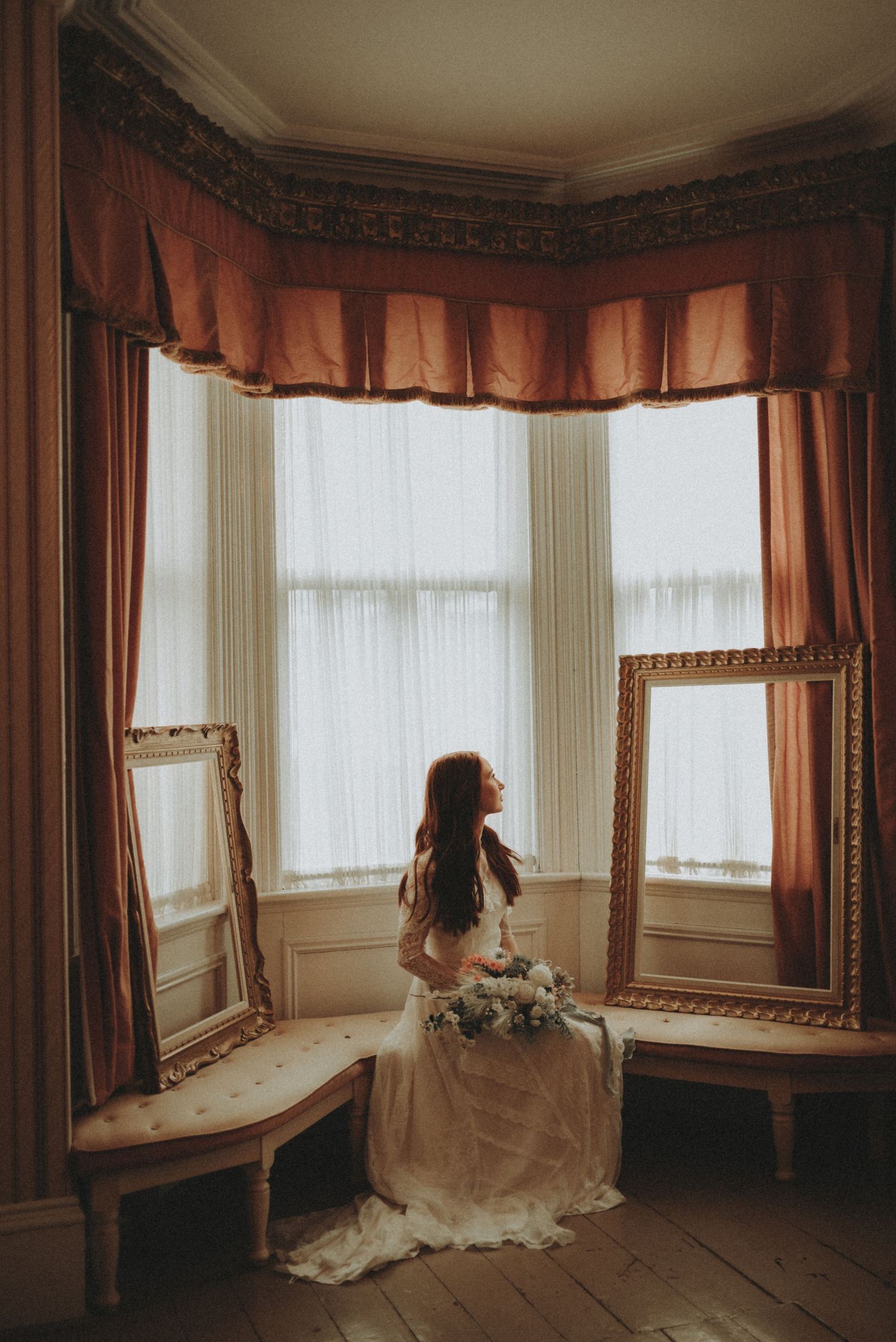Wedding dress train elegantly draped across vintage hardwood floors near bay windows and ornate curtains.