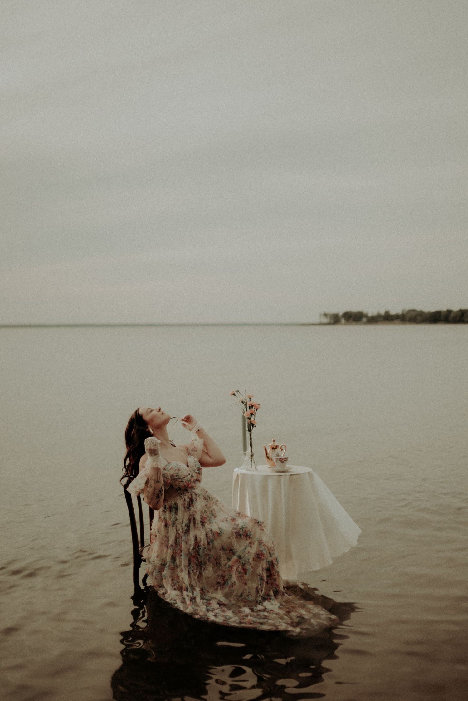 Dramatic pose in a floral gown while seated in shallow water with vintage furniture and table setting.