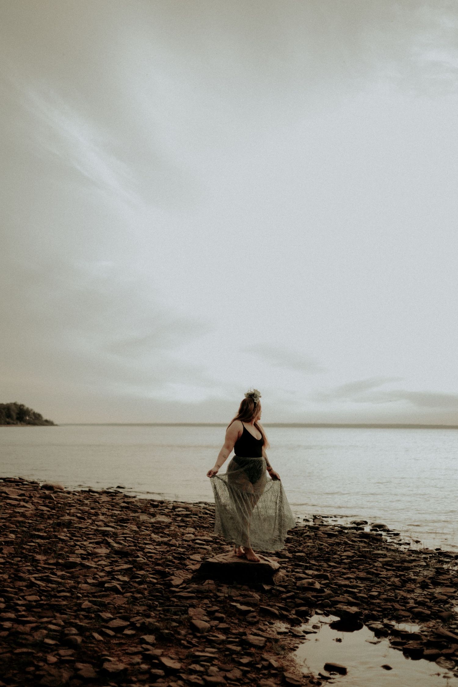 Silhouette stands on rocky beach shore with flowing dress against cloudy sky.
