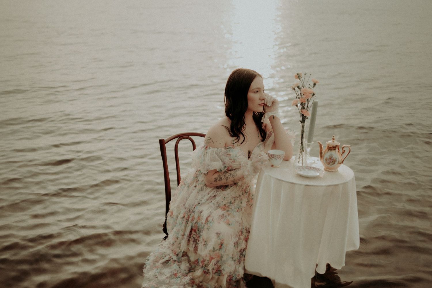 Romantic seaside scene with small table setting and floral dress against misty water backdrop.