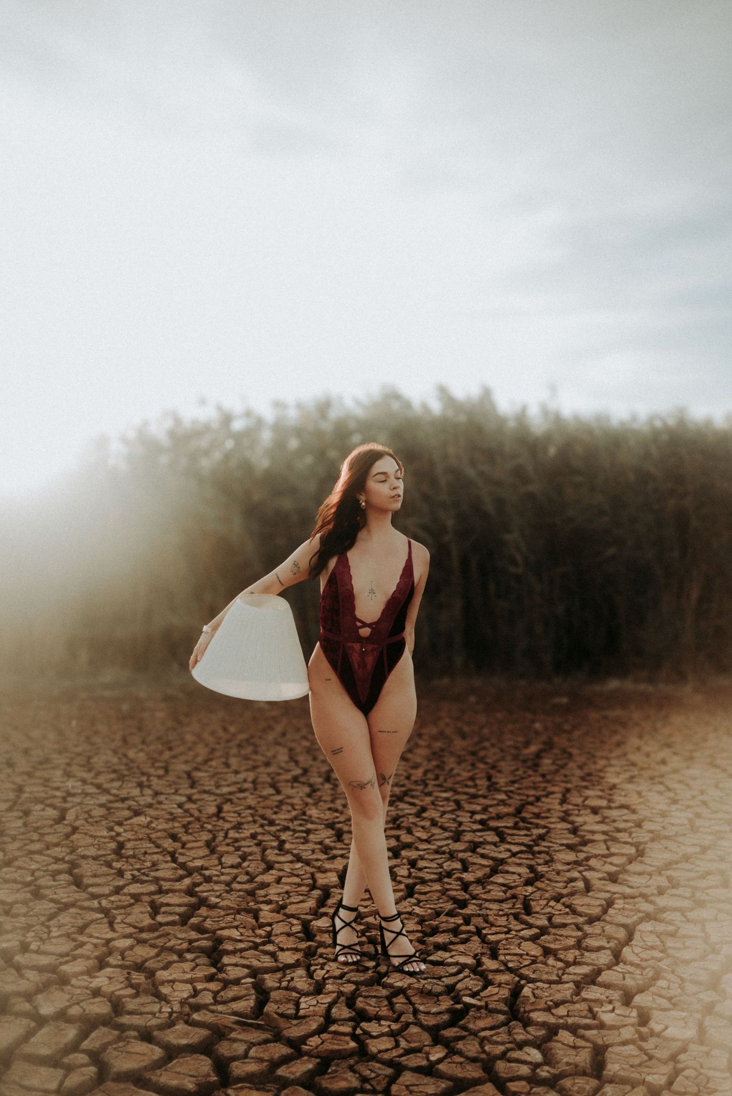 Surfer in burgundy swimsuit stands on cracked dry earth holding white surfboard at sunset.