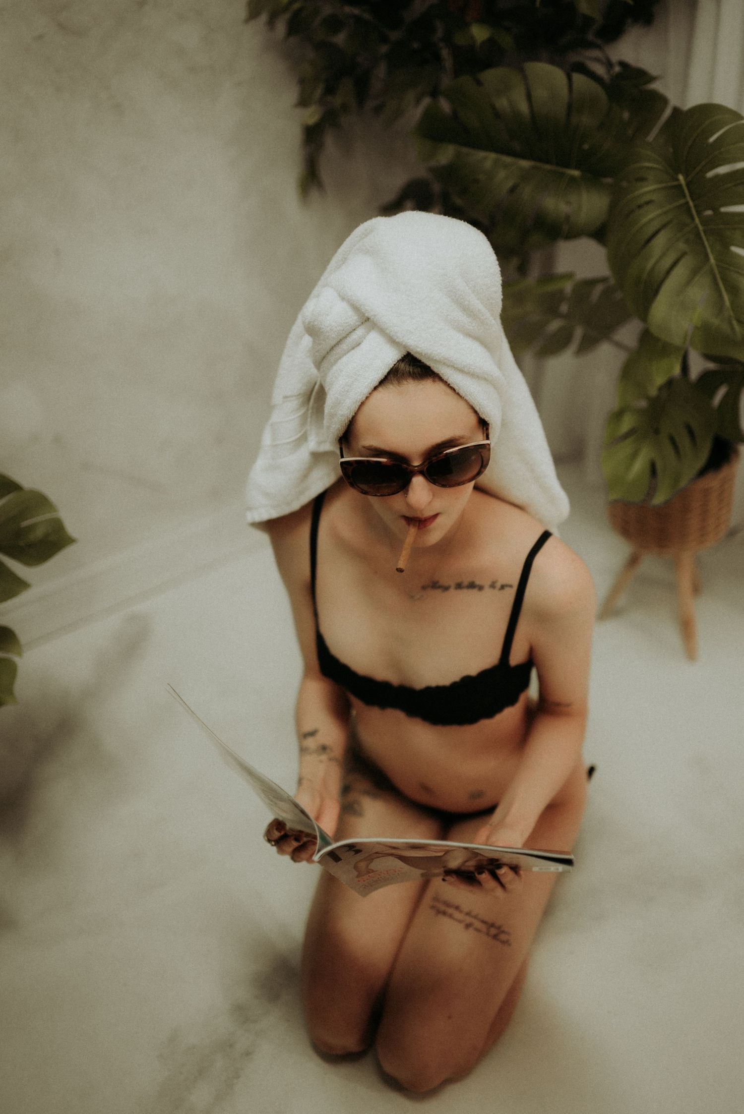 Artistic portrait with white towel turban, sunglasses and black lingerie amid tropical plants against textured wall.