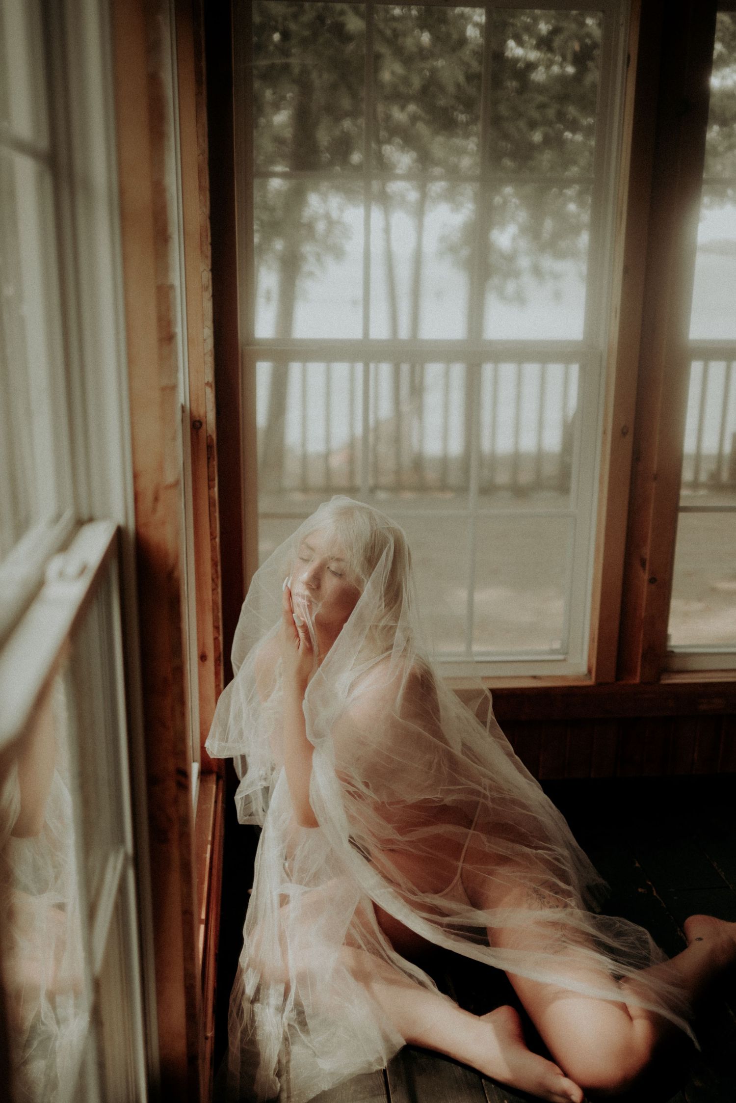 A dreamy boudoir photo featuring a sheer veil by a sunlit window in a rustic cabin.