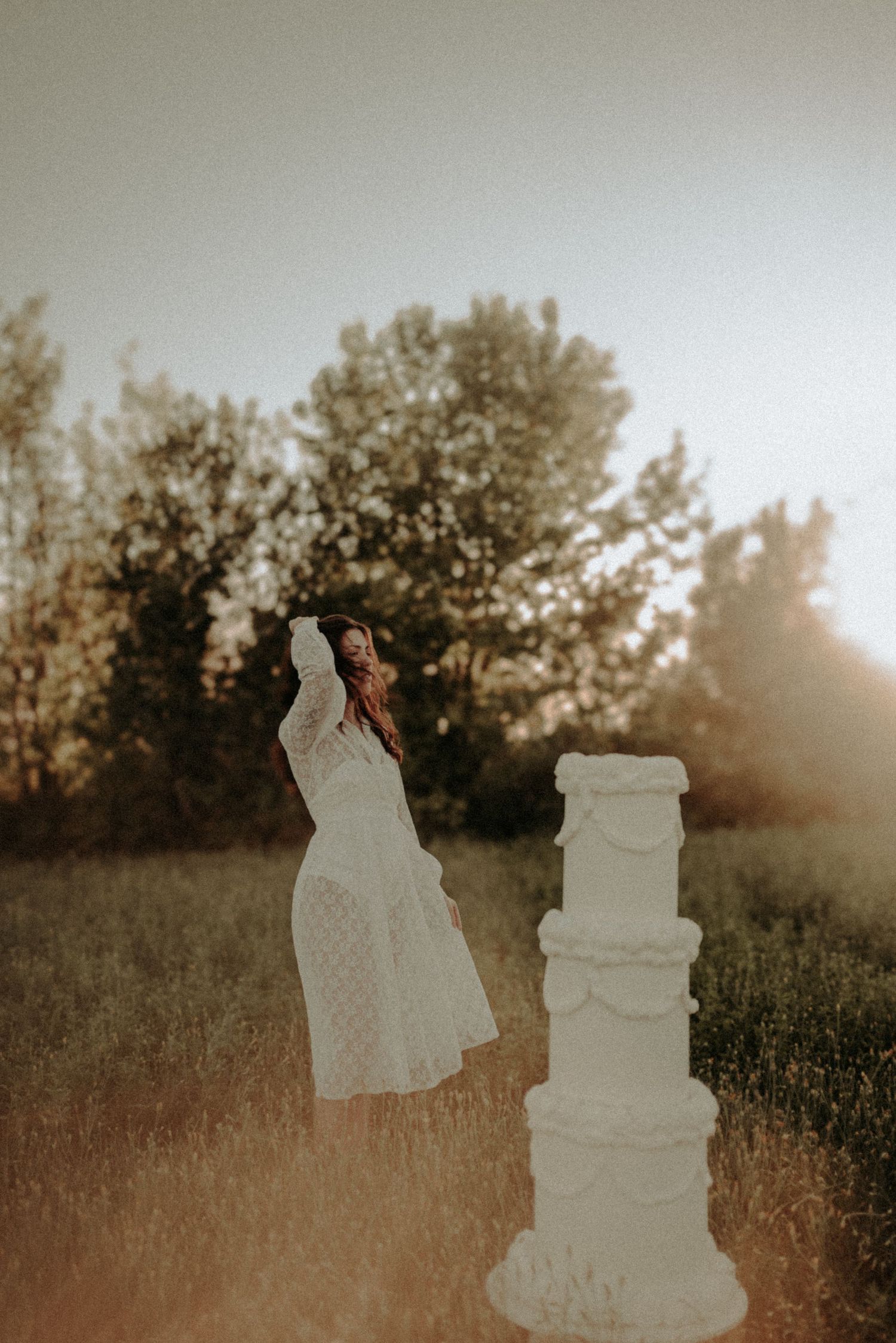 A silhouetted wedding couple stands next to their white tiered cake at sunset in a rustic outdoor setting.