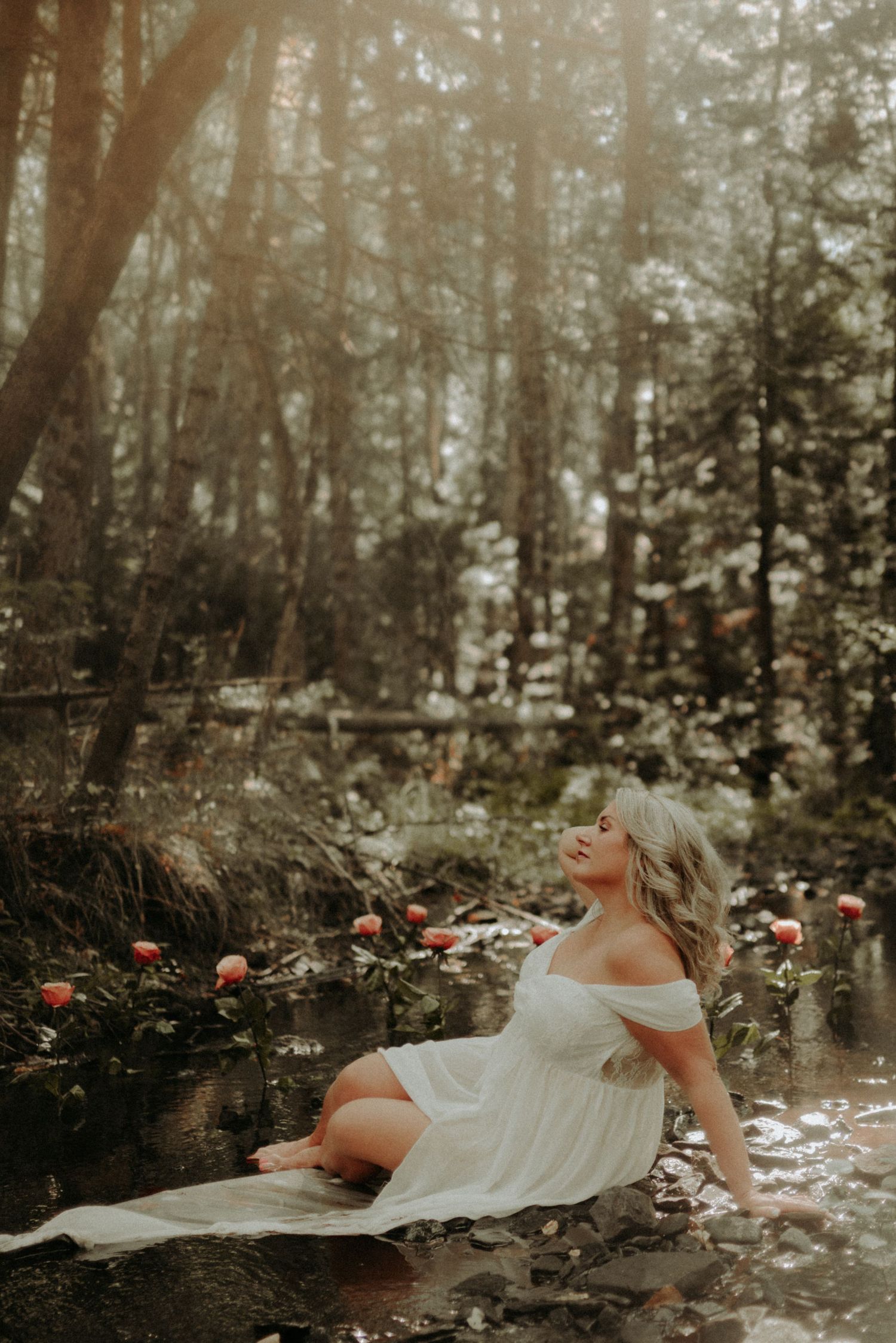 Ethereal scene of a white dress floating in dark forest water surrounded by scattered rose petals and dappled sunlight.