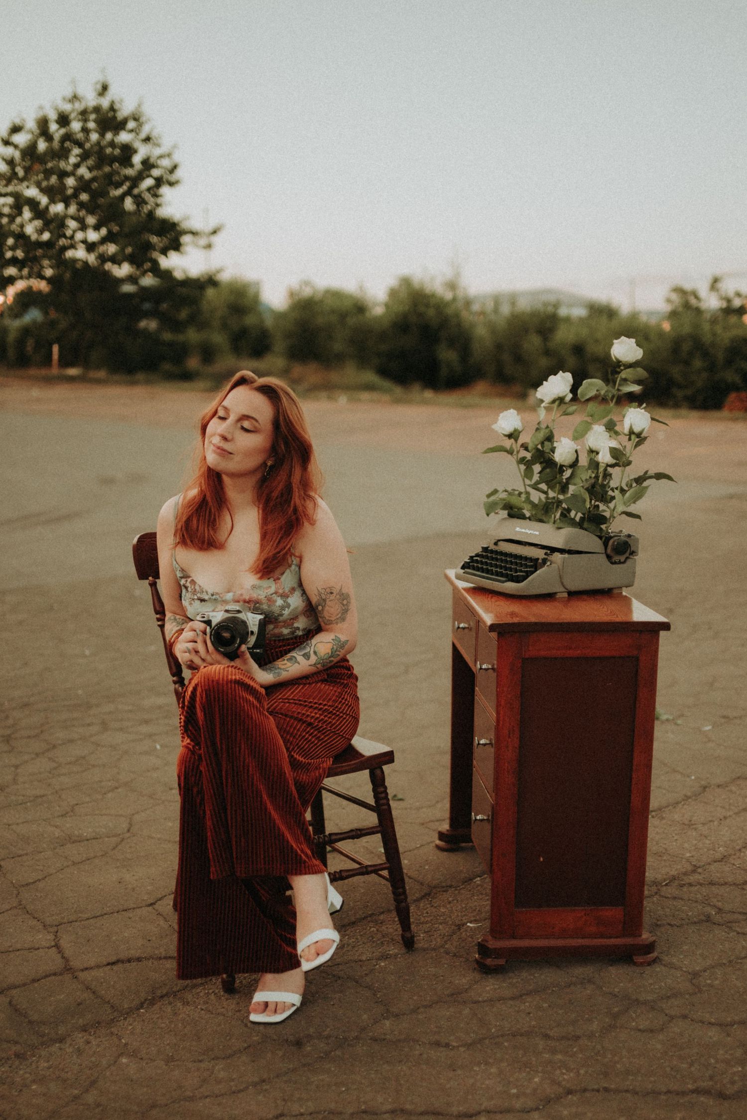 Photographer in vintage dress sits with camera near typewriter and flowers outdoors at sunset.