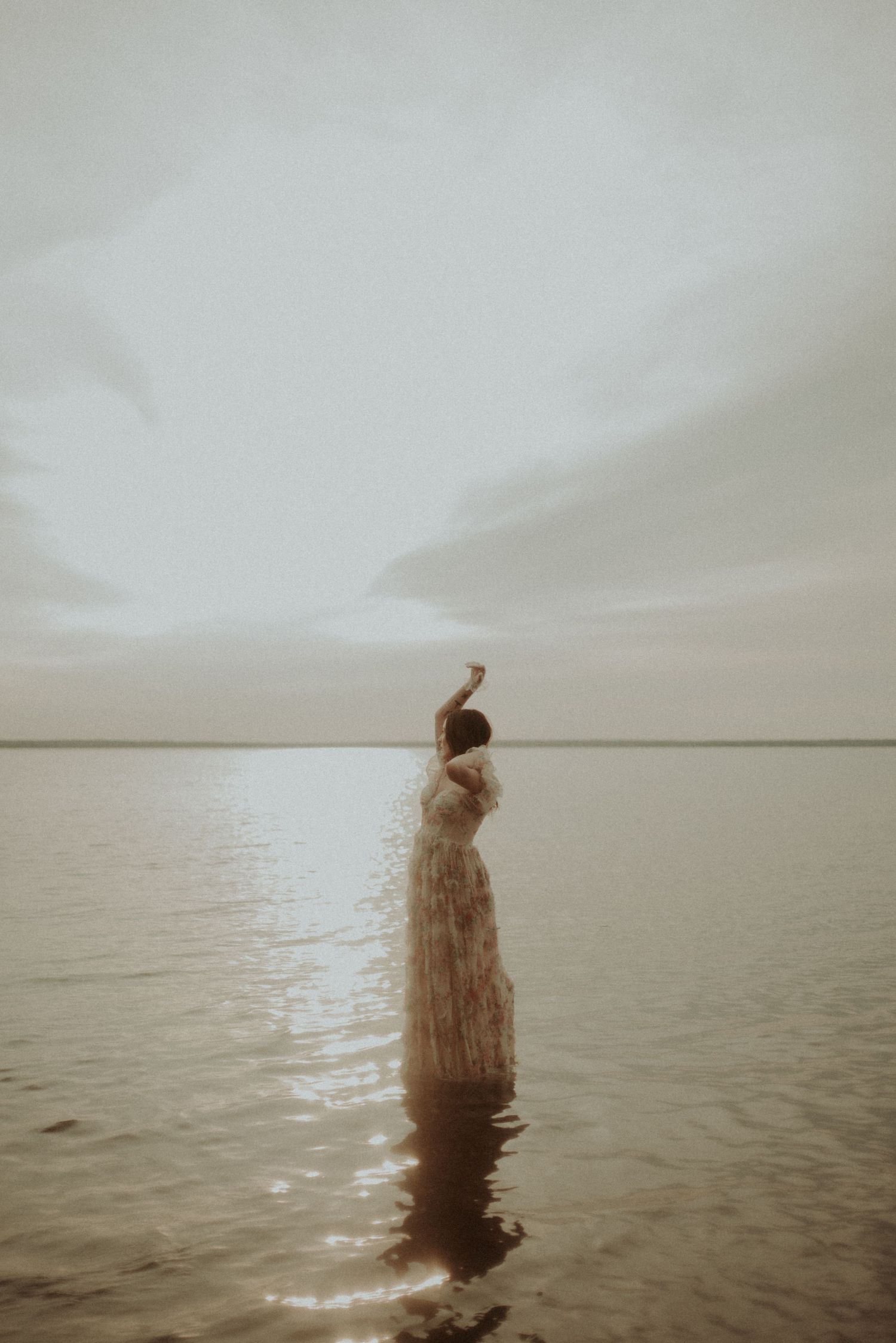 A silhouette stands in calm waters wearing a flowing dress against an overcast sky.