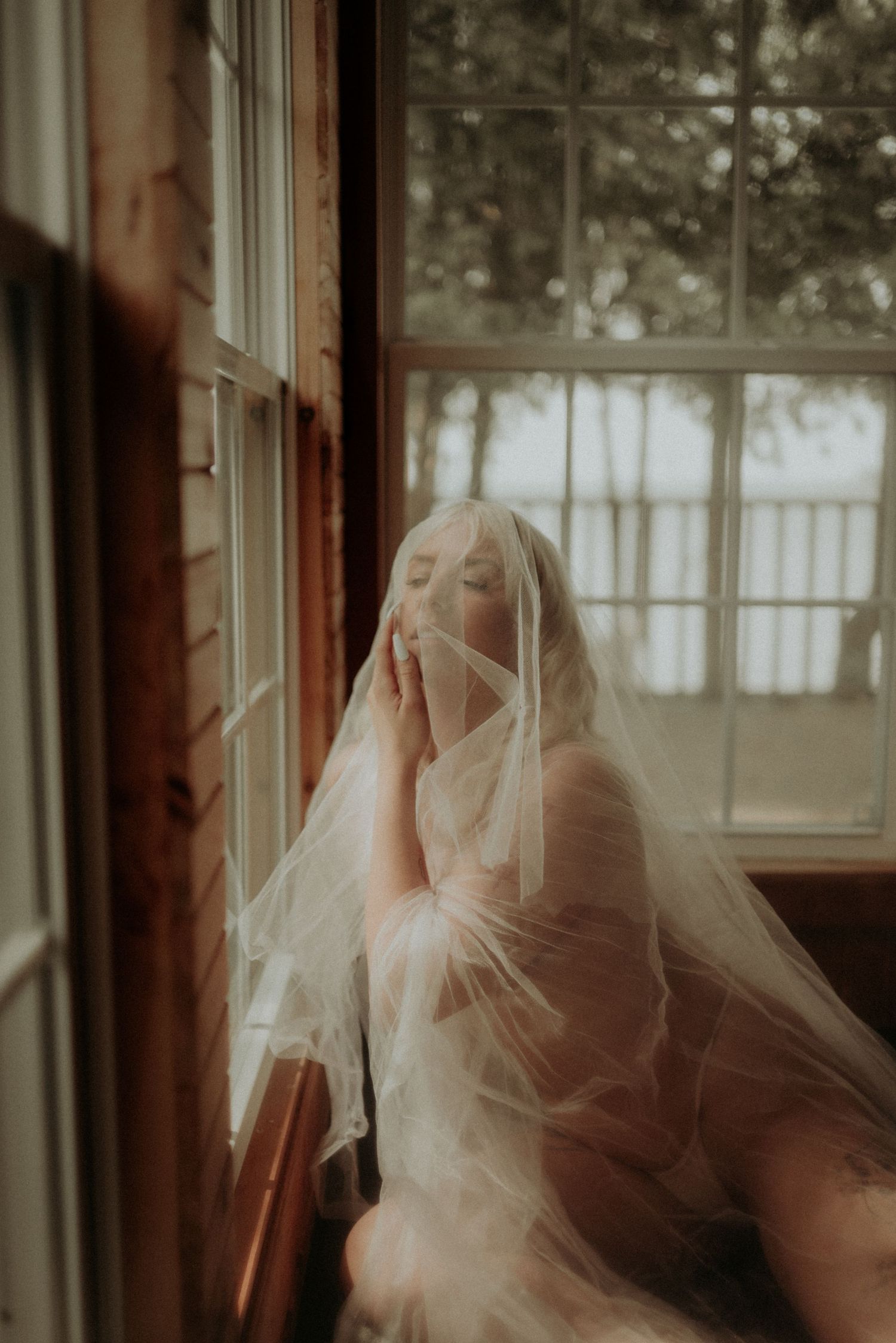 Soft and romantic boudoir photograph with translucent veil by a window.